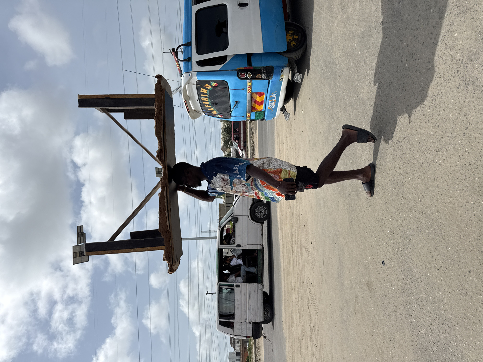 A man is walking along a paved road in Lekki, Nigeria, carrying a large, rectangular object constructed of metal and rough material on his head. He is wearing a patterned shirt and shorts, and flip-flops. In the background are two vehicles: a white van filled with people and a light-blue minibus. Overhead power lines are visible against a partly cloudy sky.