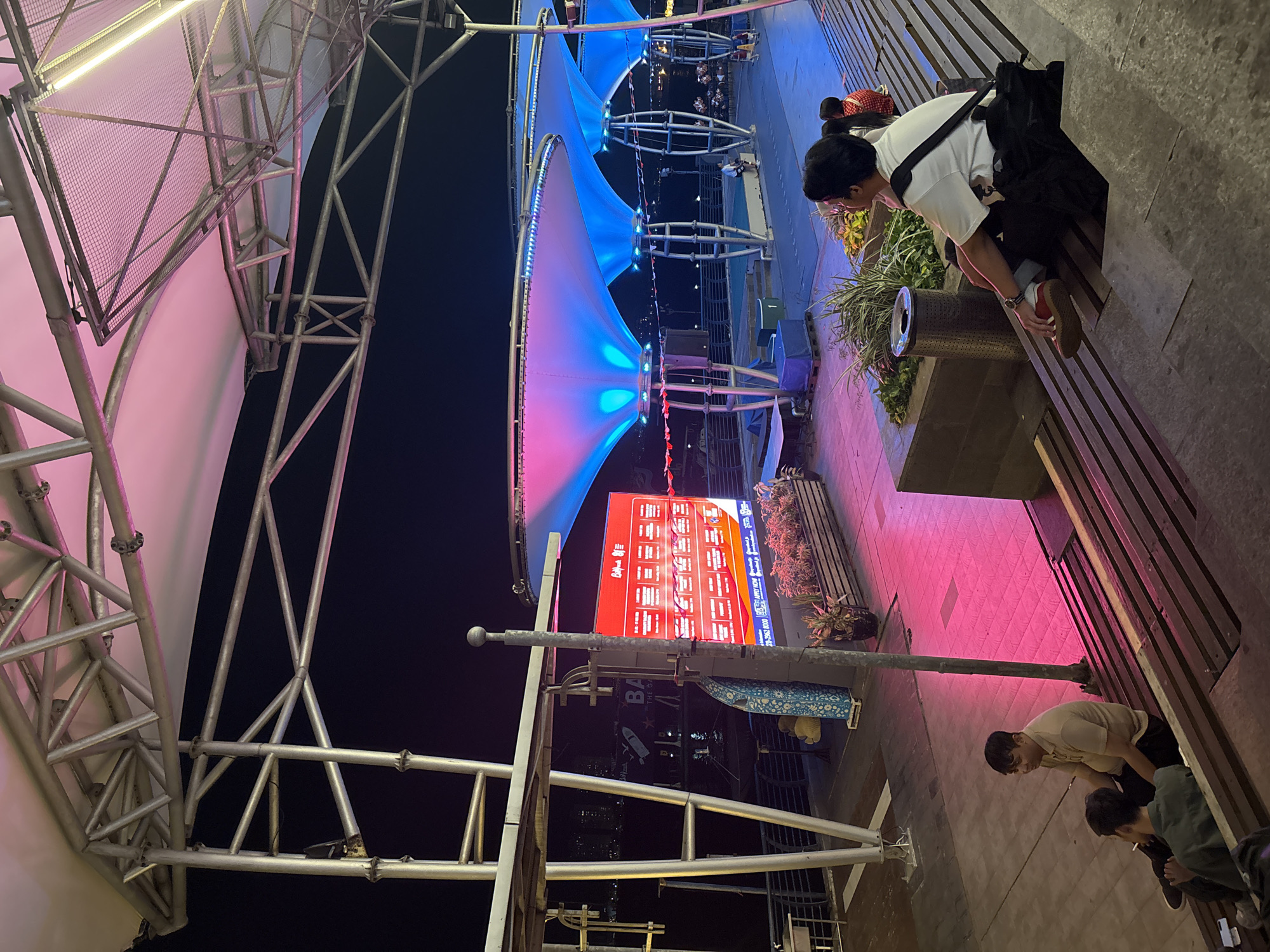 In North Jakarta, Indonesia, an outdoor seating area is illuminated by vibrant pink and blue lights from overhead canopies. A large red digital billboard displays advertisements, with the text "BA" and "THE O" visible. In the foreground, two men sit on wooden steps. One man, wearing a light brown shirt, is smoking a cigarette. Another man, wearing a white shirt and black pants, is seated with a black bag on his lap and a watch on his left wrist. Further back, a child in a red and white outfit is visible. The scene appears to be a public gathering space, possibly a waterfront promenade or entertainment district, at night.