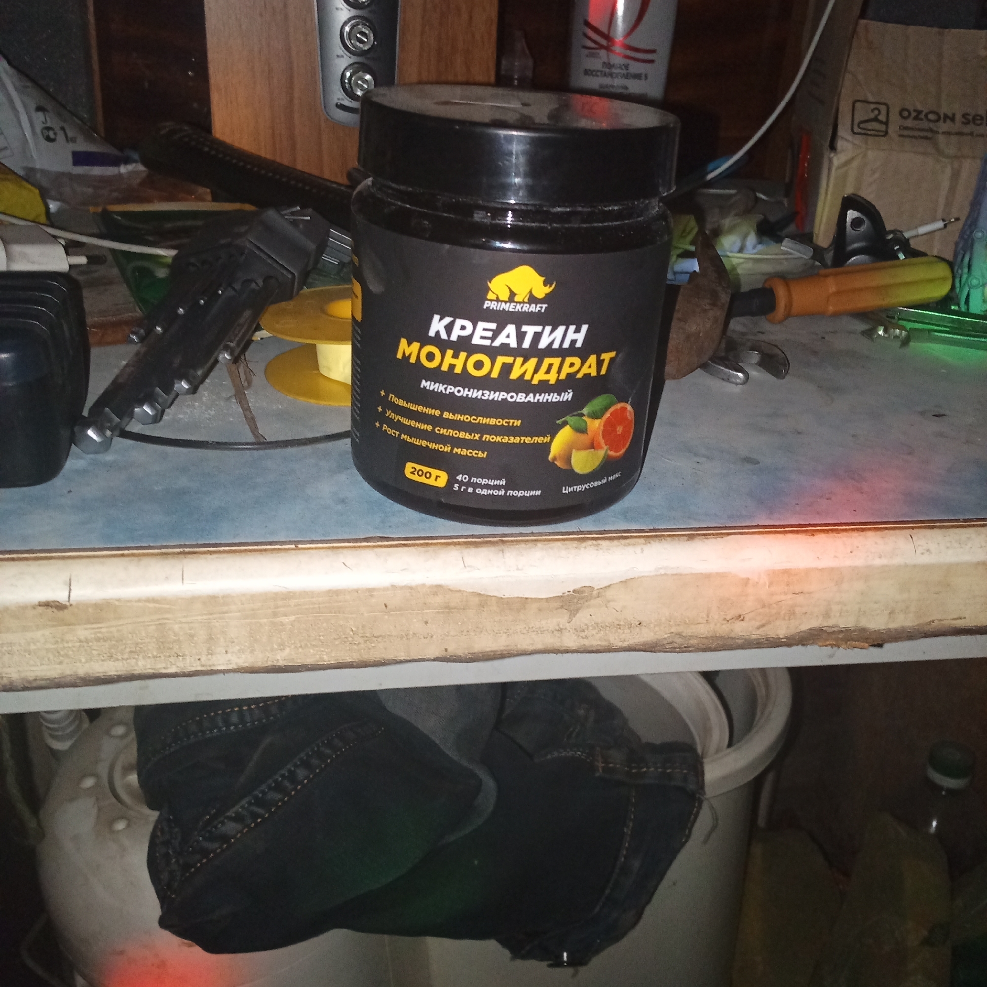 A black plastic jar containing "KREATIN MONOGIDRAT" (Creatine Monohydrate) by PRIME KRAFT with a citrus fruit illustration is placed on a shelf. The jar is predominantly black with yellow and white text. The shelf is wooden and appears to be part of a cluttered storage area. In the background, various objects are visible, including what appears to be a black tool, yellow spools, and cardboard boxes with Russian text. The lighting is dim, creating shadows and highlighting certain objects. The image does not contain any identifiable landmarks or elements that would indicate its specific location within the city of Алфёрово, Russia.