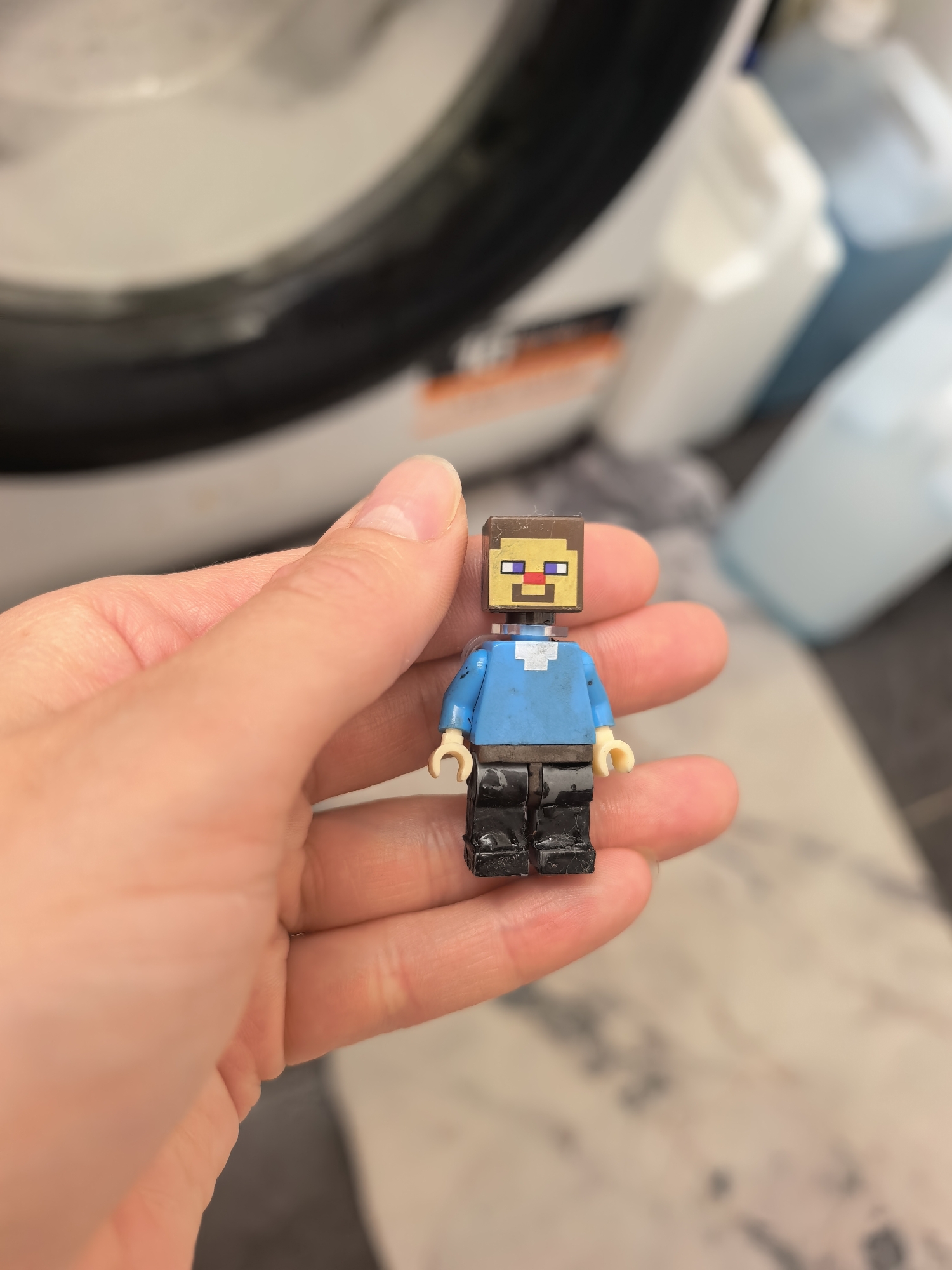 A human hand is prominently featured in the foreground, holding a LEGO minifigure in a domestic setting in Sterlitamak, Russia. The minifigure, a recognizable representation of "Steve" from the Minecraft video game, has a pixelated brown head with purple eyes and a red mouth, a blue shirt with a white collar, and black legs. The figure appears slightly worn, indicating it has been played with.
The hand, with visible skin texture and fingernails, gently cradles the small toy. The background is intentionally blurred, directing focus to the minifigure. Despite the blur, a white front-loading washing machine can be identified on the left, with its circular door seal partially visible. To the right of the washing machine, several plastic bottles, likely containing laundry products such as detergent or fabric softener, are vaguely discernible, one appearing white and another light blue.
The scene takes place indoors, likely in a laundry room or utility area, illuminated by what appears to be artificial light. There is no specific activity or event taking place other than the static pose of the hand holding the toy. No legible text is visible on any objects in the background. The image captures a simple, everyday moment in a household setting.