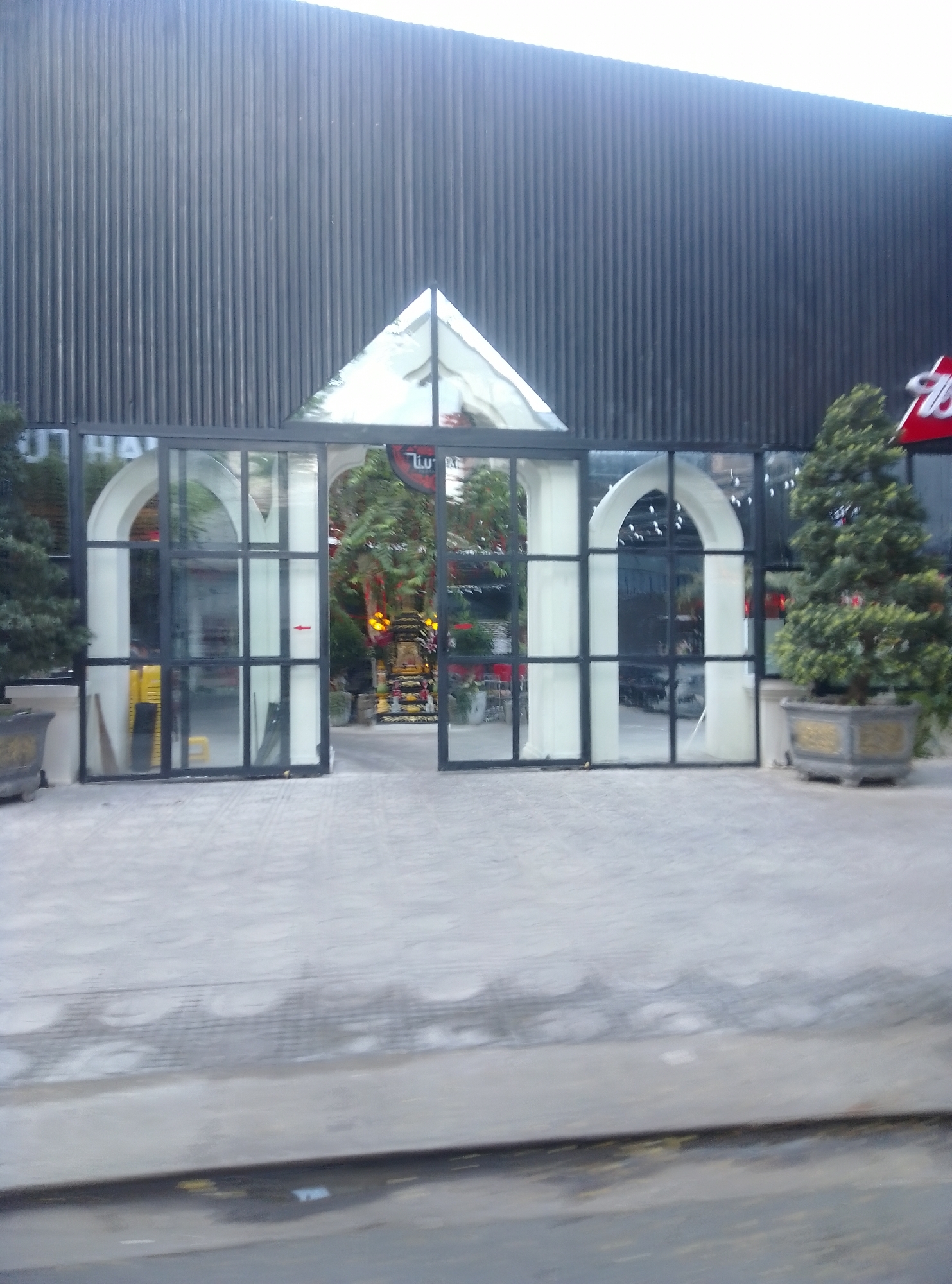 In the city of Bac Nha Trang Ward, Vietnam, a storefront features a glass facade with dark metal framing. Two arched entrances are visible through the glass. To the left, a potted evergreen tree is situated near a gray planter. To the right, another potted evergreen tree stands beside a decorative gray pot. The building's exterior wall is made of vertical dark corrugated metal. The ground in front of the building is paved with light-colored concrete. A red sign with a white graphic is visible on the right side of the building, partially obscured by the evergreen tree.