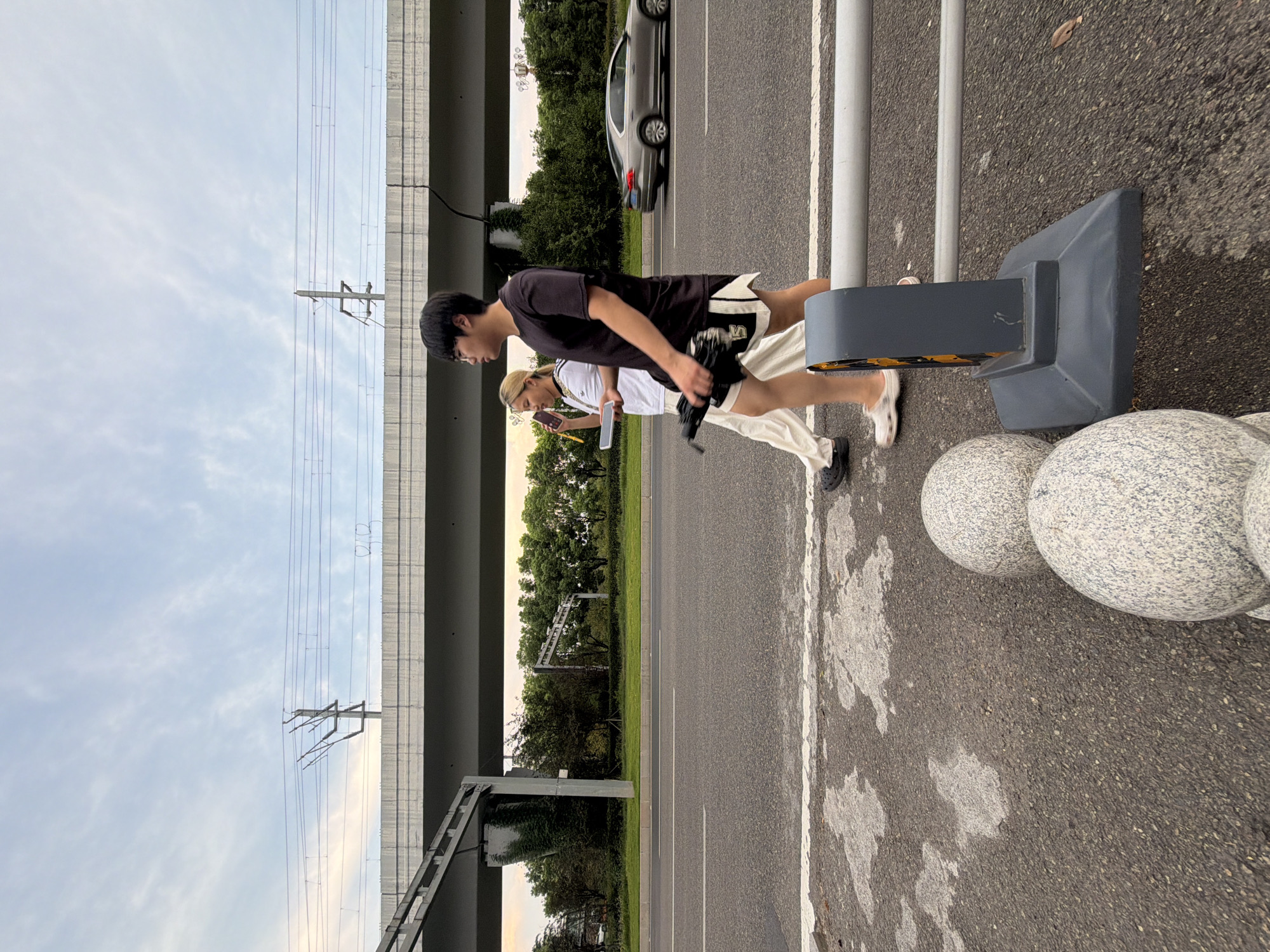 In the city of Jiangjin, China, on a road with a lane divider, two individuals are walking. The person on the left, appearing to be a woman, wears light-colored pants and a white t-shirt with black stripes. She holds a phone to her ear and a tablet in her other hand. The person on the right, appearing to be a man, wears dark shorts with white accents and a black t-shirt. He is holding a black object, possibly an umbrella, in his right hand. They are walking in the same direction, away from the camera, on the sidewalk. In the background, a silver car is driving on the road. Above the road, a raised concrete structure, possibly an overpass or bridge, supports overhead power lines. The sky is partly cloudy with a light blue hue. The foreground shows a paved surface with a white line and decorative spherical concrete bollards.