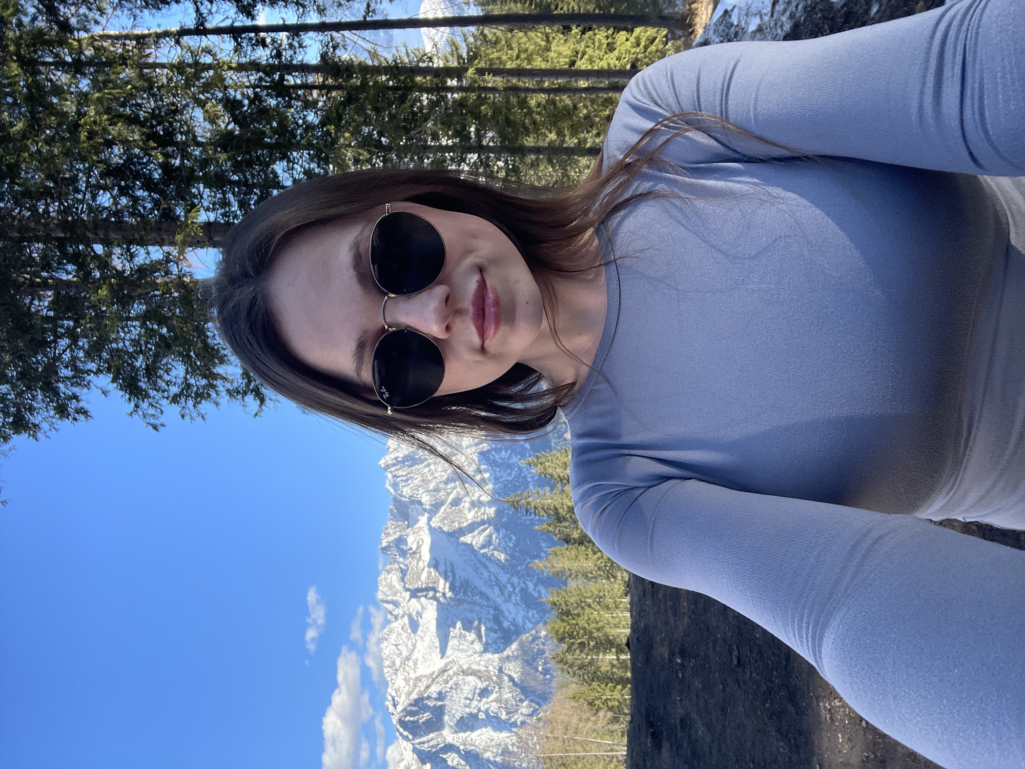 A woman with long brown hair and sunglasses poses for a selfie outdoors. Behind her is a landscape featuring snow-capped mountains, evergreen trees, and a clear blue sky. Based on the mountainous terrain, the scene is possibly in Cortina d'Ampezzo, Italy. The woman is wearing a light blue, form-fitting long-sleeved shirt.