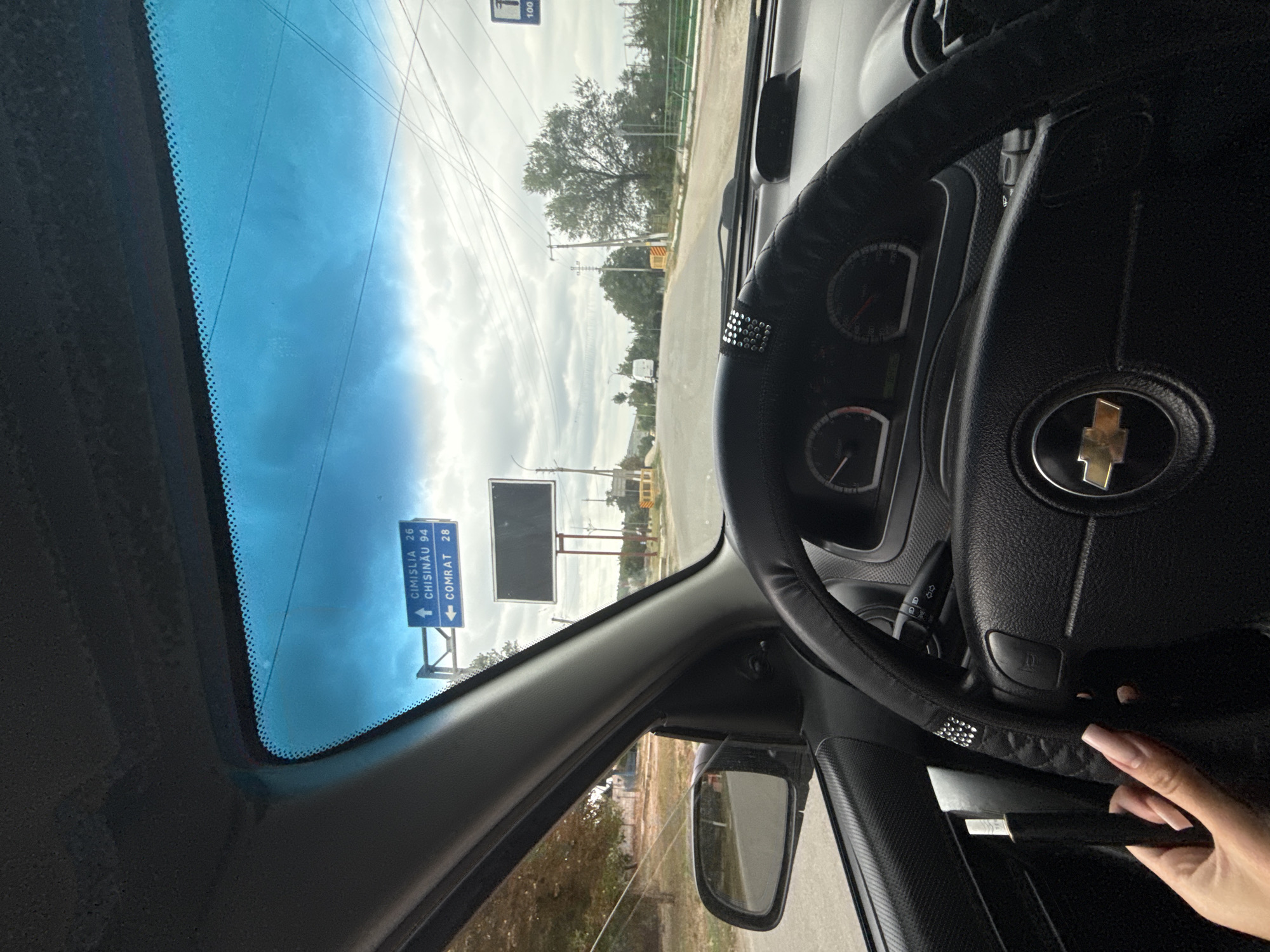 The image is taken from inside a car, looking forward through the windshield. The dashboard and steering wheel are visible in the foreground, with a hand with long, manicured nails holding a black object. A road with signs is visible in the distance, under a cloudy sky. One sign indicates directions to Cimislia (26 km) and Chisinau (94 km) with an upward arrow, and to Comrat (28 km) with a leftward arrow. This signage suggests the location is on a road leading out of or towards Basarabeasca, Moldova, given the proximity of these cities. A large, blank billboard is also present near the road. The environment appears to be semi-rural, with trees and utility poles visible along the roadside.