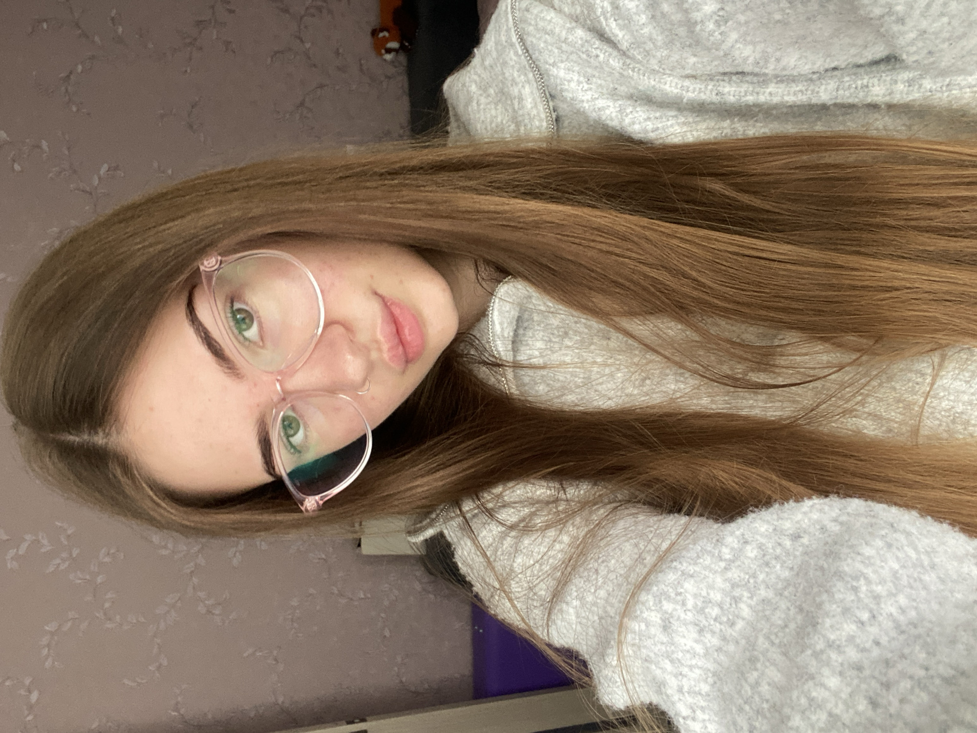 A close-up, angled portrait features a young person with long, straight, reddish-brown hair. They are looking directly towards the camera, wearing light pink or clear-framed glasses that emphasize their green eyes. Their lips are slightly parted, and their expression is neutral and relaxed. The person is dressed in a light gray, textured hooded sweatshirt with a visible zipper down the front.
The background consists of a wall covered in light brownish-pink or mauve wallpaper adorned with a subtle, repeating pattern of leaves or vines. In the upper left corner of the background, a dark object is partially visible, and a purple item can be seen in the lower right. The lighting is soft and even, indicating an indoor setting. There is no discernible text in the image.