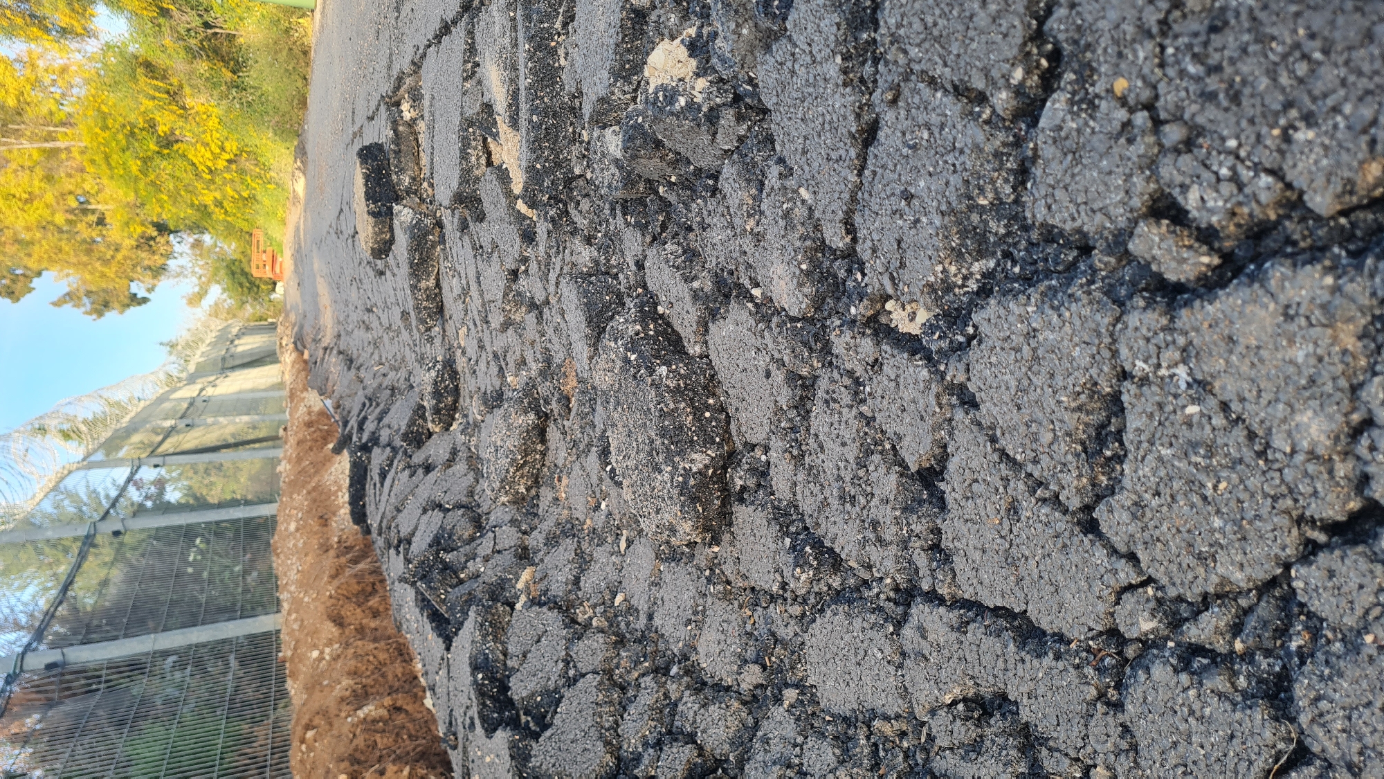 The image shows a severely damaged asphalt road in the Lachish Regional Council, Israel. Extensive cracking and crumbling of the asphalt are visible. Adjacent to the road is a dirt shoulder and a high security fence with razor wire. In the background, there are trees and a blue sky.