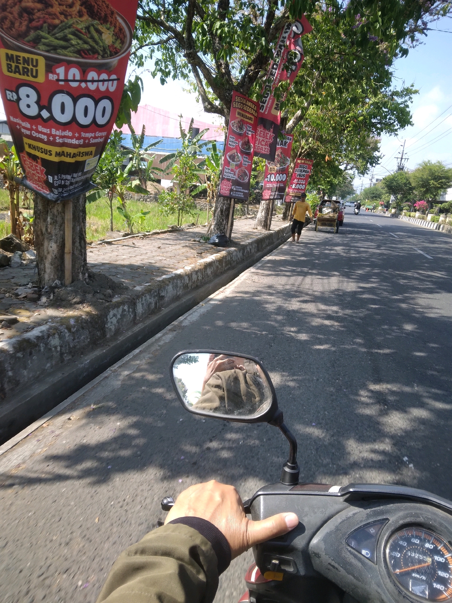 The image shows a first-person perspective of someone riding a scooter on a street in Purwokerto Kulon, Indonesia. The rider's hand is visible on the handlebars, and the scooter's speedometer is in the lower right corner. The street is paved and lined with trees casting shadows on the road. On the left side of the road, there are banners advertising food specials, with prices and menu items visible, including "MENU BARU RP 10.000 RP 8.000". A person is visible pushing a cart on the side of the road. In the background, there are buildings and other vehicles, including a motorcycle. The overall scene depicts a typical street view in an Indonesian city.