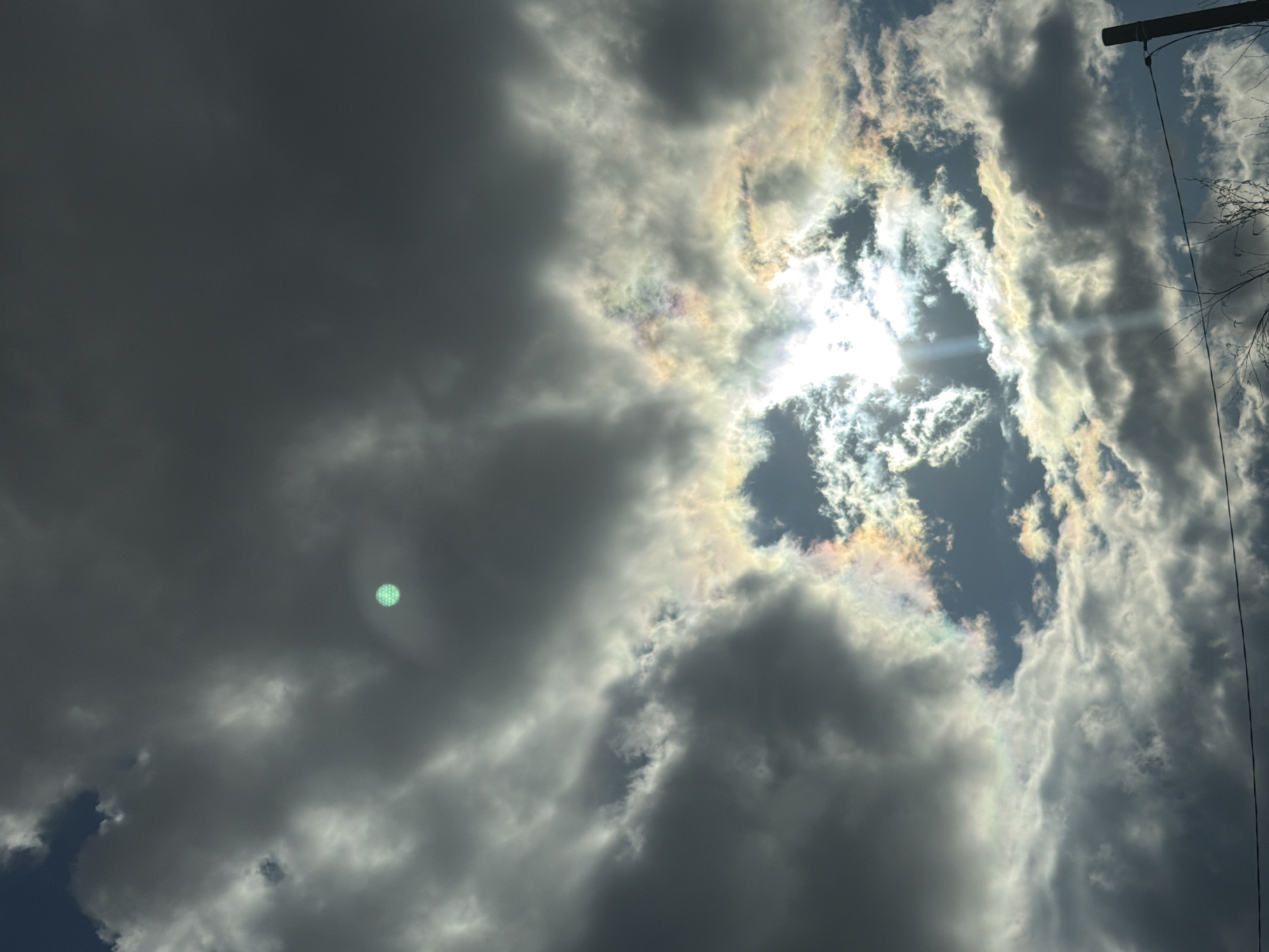 The image shows a daytime sky with dramatic cloud formations. A bright sun is visible through a break in the clouds, which are dark gray and white. Iridescent colors appear around the edge of some clouds. A power line and some bare tree branches are visible in the upper right corner of the frame. The image was taken looking upwards. There is nothing in the photograph to confirm the location is in Chaikovsky, Russia.