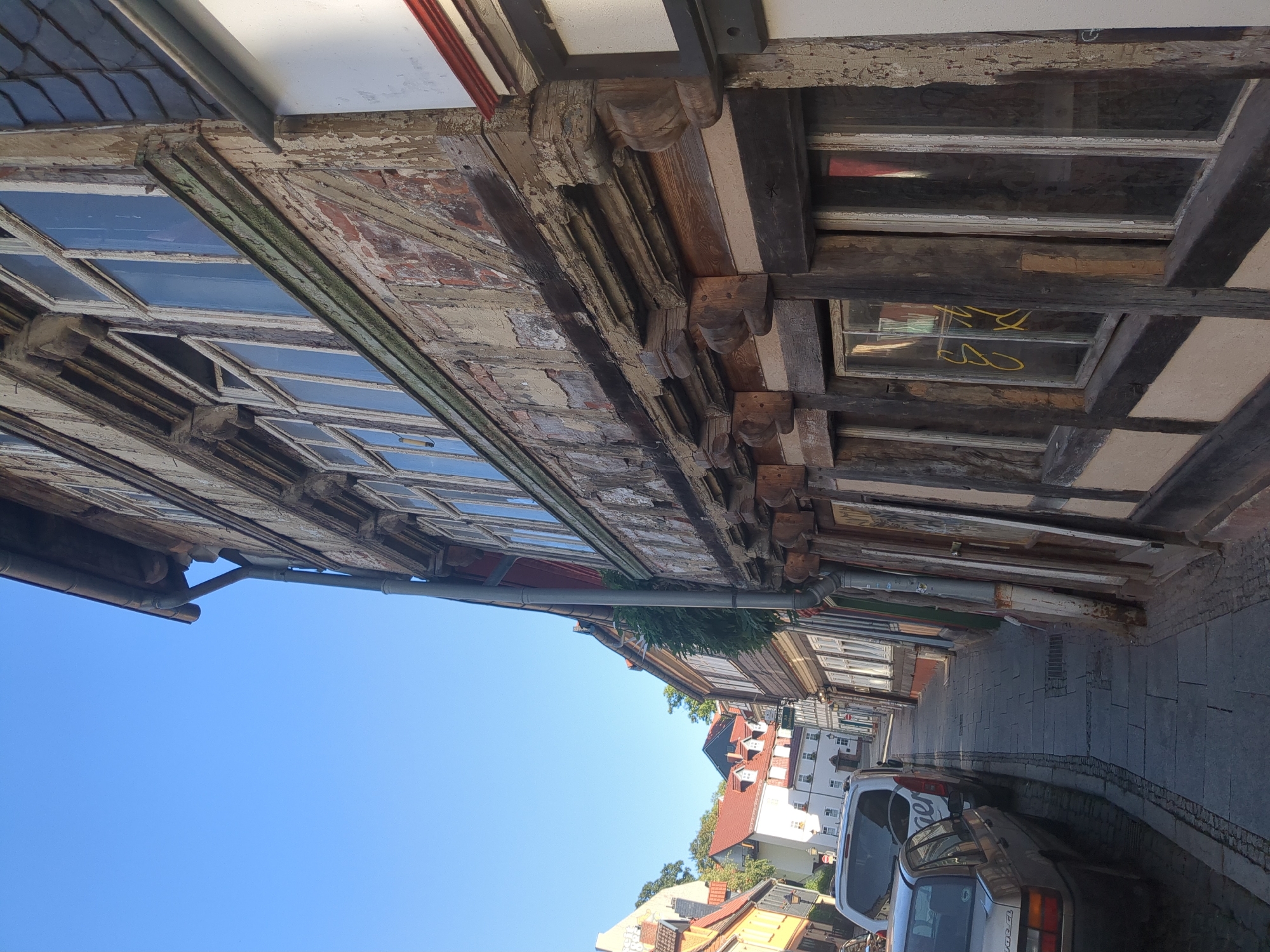 The image captures a perspective looking down a narrow cobblestone street in Nordhausen, Germany. The street is flanked on the right by an old, timber-framed building with weathered wooden beams and plaster walls, showing signs of age and peeling paint. The building features numerous multi-paned windows. On the left side of the street, parked cars are visible, including a silver sedan and a white van. The sky above is clear and blue. Further down the street, other historical buildings and trees can be seen, contributing to the ambiance of an old European town.