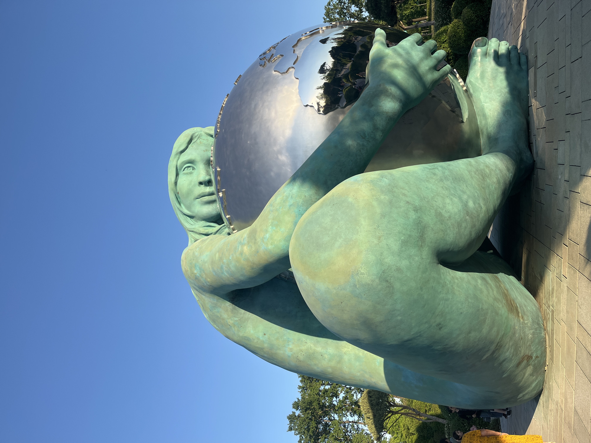 A large, imposing statue of a woman made from a green-patinated material, resembling weathered bronze or copper, is the central subject of the image. The woman is depicted in a crouched or seated position, embracing and supporting a large, highly reflective metallic globe with both hands and arms. Her facial features are serene and contemplative, with her gaze directed towards the globe. Her hair is styled in soft, sculpted waves.
The globe is smooth and shiny, with a silver-like finish that acts as a mirror, reflecting the clear blue sky, fluffy white clouds, and distant trees. Sculpted landmasses, representing continents, are visible on its surface. The statue itself shows a range of green tones with some darker, brownish streaks, characteristic of aged bronze. Detailed sculpting is evident in the woman's hands and feet, which are firmly planted on the ground.
The setting is outdoors, under a vibrant, cloudless blue sky. Bright sunlight illuminates the scene, creating distinct shadows on the statue and the ground, suggesting it is a clear, sunny day. The statue rests on light-colored, block-patterned paving stones. In the background, green foliage from trees and shrubs is visible, along with glimpses of architectural elements or surrounding landscape. A few people are faintly visible near the base of the statue in the full image, providing an indication of its large scale. No visible text is present in the image. The location of this statue is Krasnodar, Russia.
