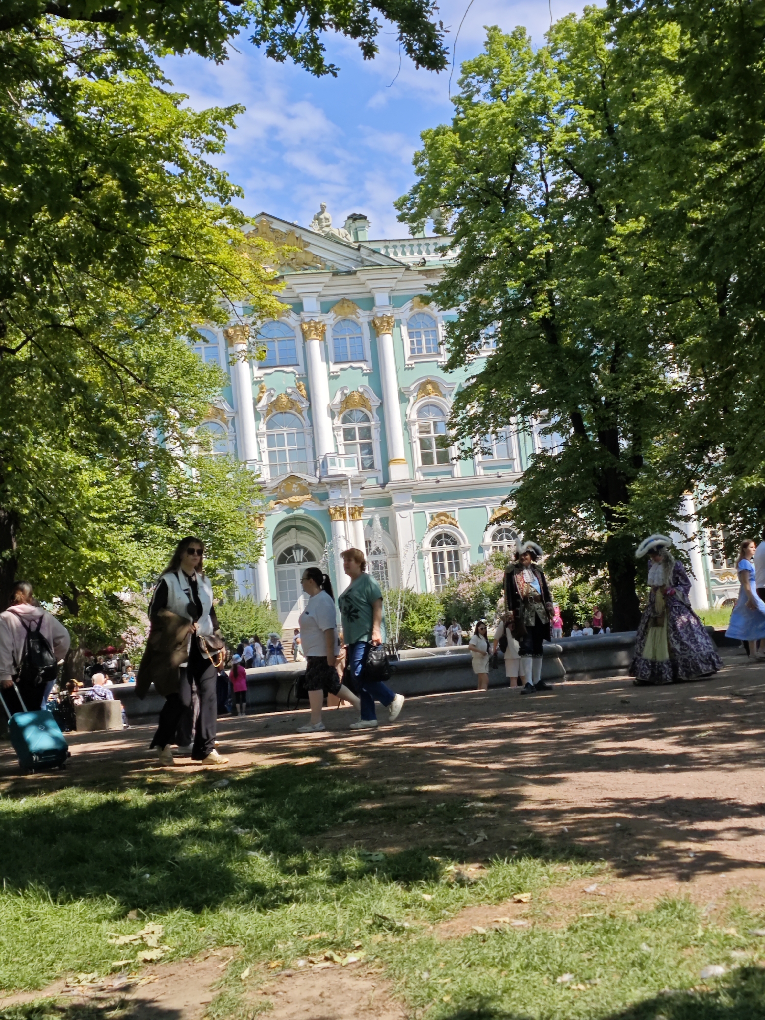 This image depicts the Winter Palace in Saint Petersburg, Russia, a grand Baroque building with a pale turquoise facade and white and gold detailing. The palace is framed by lush green trees, with dappled sunlight casting shadows on the ground. Several people are visible in the foreground and middle ground. On the left, a woman with dark hair and sunglasses, wearing a white vest over a dark top, walks with a brown bag slung over her shoulder. She is followed by a person pulling a blue suitcase. Further to the right, several individuals are walking, including two women in casual attire and two people dressed in historical costumes – one man in a dark outfit with a feathered hat and a woman in a long, ornate dress. In the background, a fountain is visible, and more people are gathered near the palace. The scene appears to be set on a sunny day in what could be a park or public square adjacent to the palace.