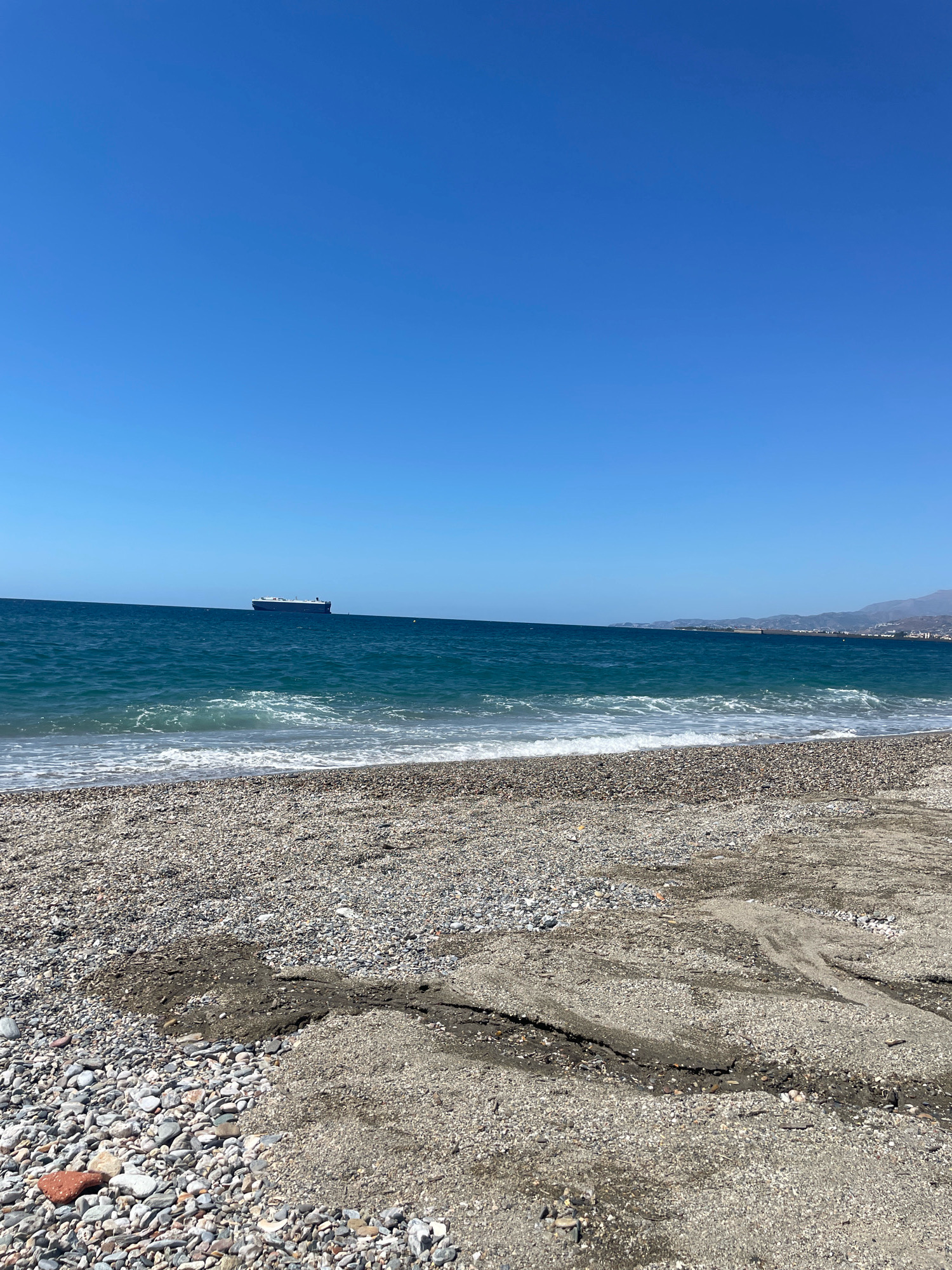 The image captures a wide view of a tranquil beach in Torrenueva Costa, Spain. The foreground is dominated by a pebble-strewn shore, with a patch of sand showing intricate patterns of dried mud and cracks. The sea, a vibrant shade of blue, stretches to the horizon, where gentle waves break along the shore, leaving white foam. In the distance, a large cargo ship is visible on the calm waters. The sky is a clear, cloudless blue, and a faint outline of hills can be seen along the coastline to the right. The scene is devoid of any people, suggesting a peaceful, unpopulated moment on the coast.