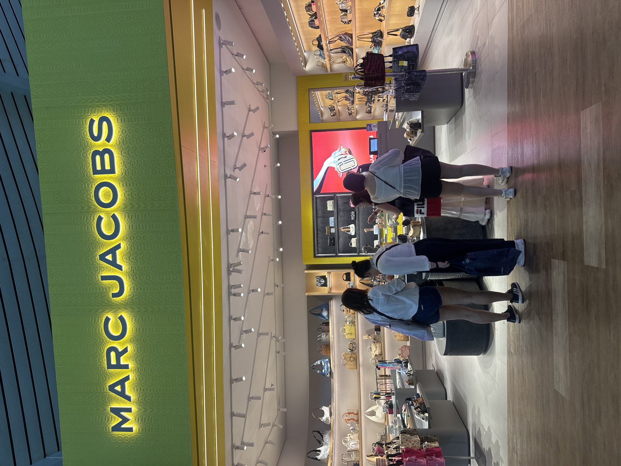The image shows the interior of a Marc Jacobs store. The "Marc Jacobs" sign is vertically oriented, lit up in yellow. Inside the store, three women are browsing through the items on display. The store has a variety of bags and accessories on shelves and display cases. The location is likely in Kuta, Indonesia.