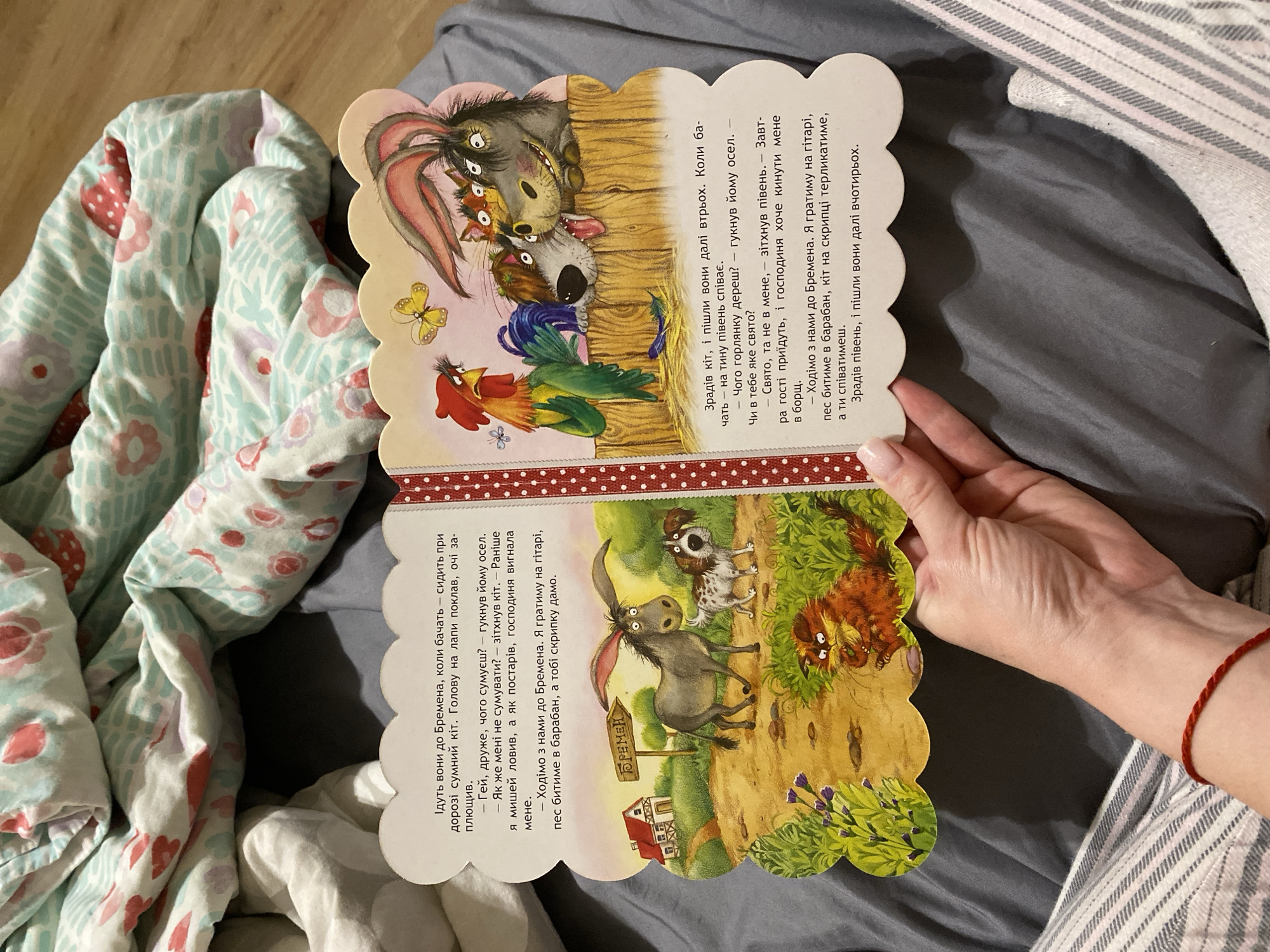 This image shows a woman's hand holding an open children's book. The book is illustrated with anthropomorphic animals. On the left page, there is text in Ukrainian and a drawing of a donkey and a dog standing on a dirt path with a house and a sign that reads "БРЕМЕН" in the background. On the right page, there is more Ukrainian text and a drawing of a rooster, three dogs, and a rabbit-like creature gathered around a wooden fence. The book is lying on a bed with gray sheets and a colorful, patterned blanket. There is no indication in the image that it is located in Kryvyi Rih, Ukraine.