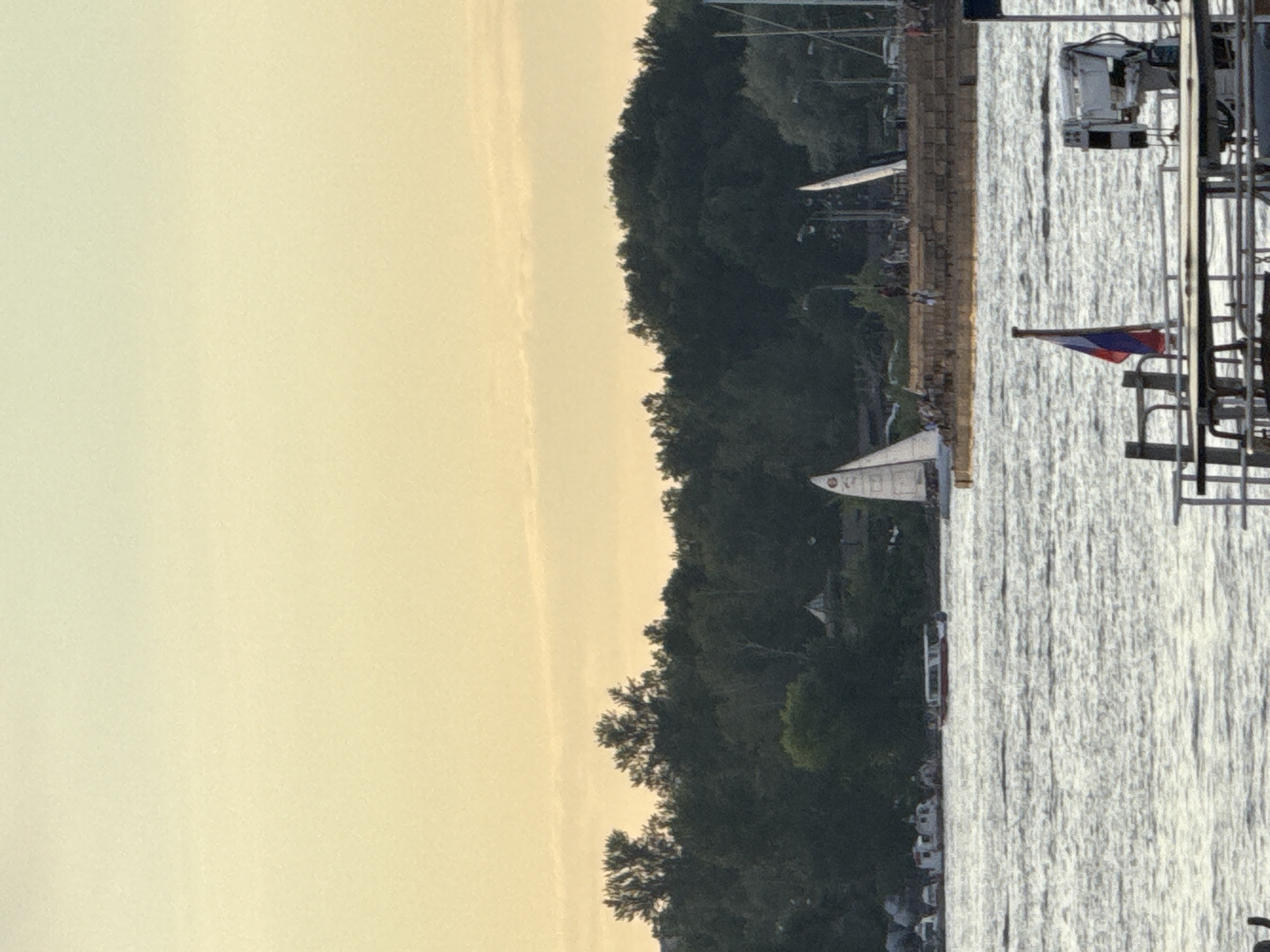 The image presents a serene waterside vista, likely captured during either sunrise or sunset, given the warm, pale yellow-orange glow that dominates the sky. The scene is divided horizontally by a body of water in the foreground and a strip of densely forested land in the middle ground, separated by a distinct embankment.
On the left side of the image, a lush expanse of green trees forms a natural, verdant backdrop, suggesting a park or forested area lining the shore. Hints of structures or buildings are nestled among the foliage. The right side features a calm body of water, possibly a river or canal, reflecting the sunlight in shimmering patterns across its surface.
A sturdy embankment, appearing to be made of stone or concrete, runs along the water's edge, forming a clear demarcation between land and water. Several boats dot the waterway and the shoreline. A white sailboat with its mast clearly visible is positioned on the water closer to the embankment. Further along the bank, other vessels are docked, including a larger white boat with superstructure visible.
A prominent detail is a small flag, displaying the red, blue, and white colors of the Russian tricolor, flown from a pole near the water's edge on the right. This, along with the given location context, firmly places the scene in Saint Petersburg, Russia. Though indistinct due to distance, small figures can be observed on the embankment, suggesting the presence of people enjoying the tranquil atmosphere. No visible text is present in the image.