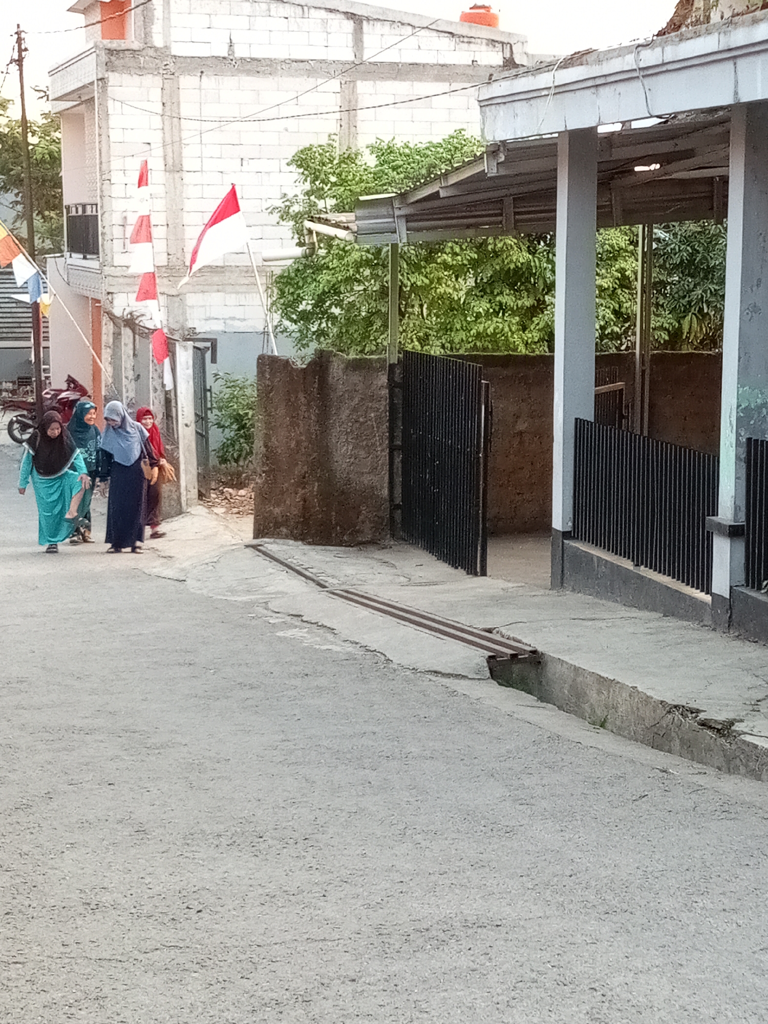 In the city of Cihanjuang, Indonesia, a paved street runs alongside a weathered building. On the left, four women are walking, each wearing a hijab and long clothing. The woman in the turquoise hijab is slightly ahead of the others. To the right of the street, a corrugated metal roof extends from the building, supported by grey pillars, with a black metal fence running along its perimeter. A drainage ditch runs parallel to the street in the foreground. In the background, a building under construction with white brick walls is visible, adorned with Indonesian flags, suggesting a national holiday or celebration. Trees with lush green foliage are interspersed between the structures.