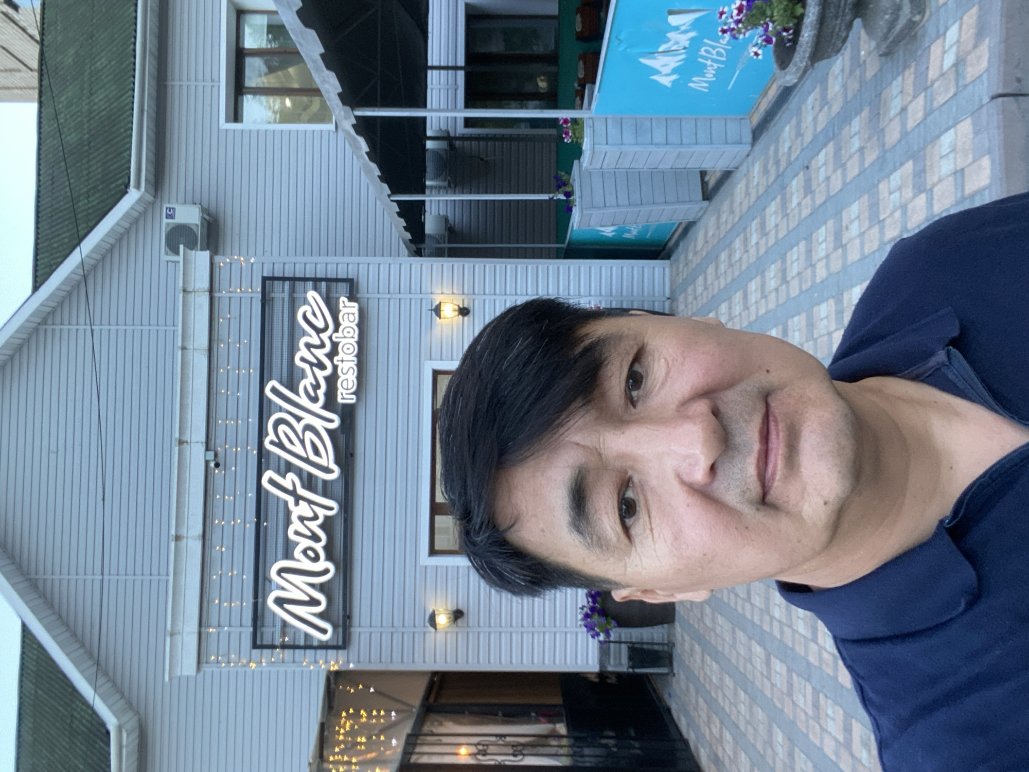 A man is standing in front of a building with a sign that reads "Mont Blanc restobar" in Petropavl, Kazakhstan. The man is wearing a dark blue polo shirt and has black hair. The building is light gray with white trim. There is a green roof and a green awning over the entrance. The ground in front of the building is paved with bricks. There are potted plants on either side of the entrance. The photo appears to have been taken in the late afternoon or early evening, as the lights on the sign are illuminated.