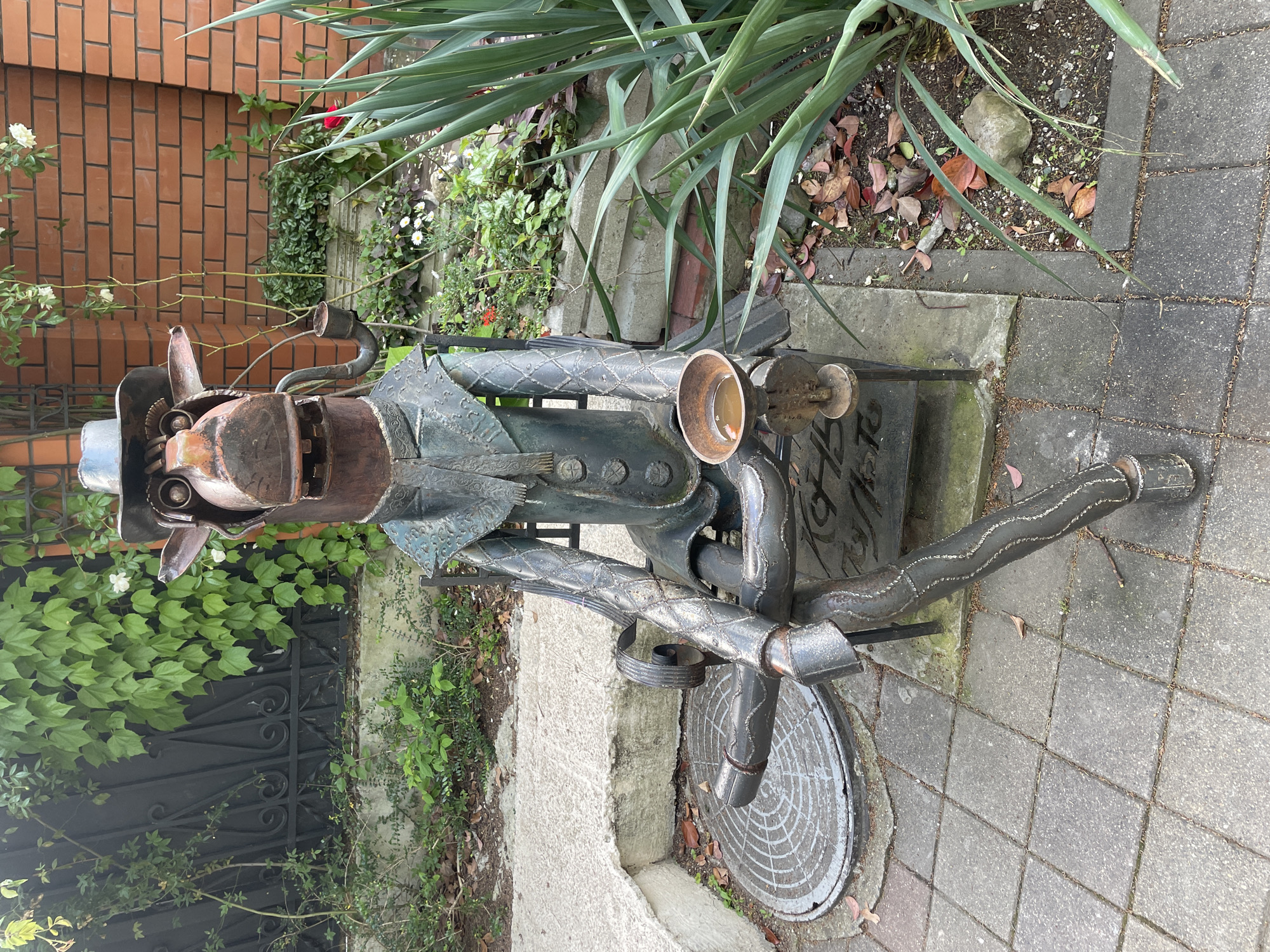 A metal sculpture of a man in a hat sitting on a bench is situated on a paved area near greenery in Sochi, Russia. The sculpture appears to be made from welded metal pieces. The background includes a brick wall and a variety of plants. A manhole cover is visible near the sculpture.