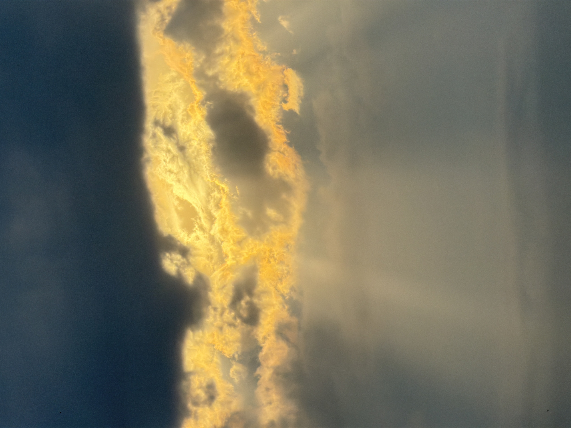 The image shows a vertical view of the sky in Tbilisi, Georgia. On the left is a large, dark mass of clouds. To the right of the dark clouds, there is a band of bright, golden-yellow clouds, possibly illuminated by the setting or rising sun. Further to the right, the sky is a muted gray-blue color, with faint beams of light shining through the clouds.