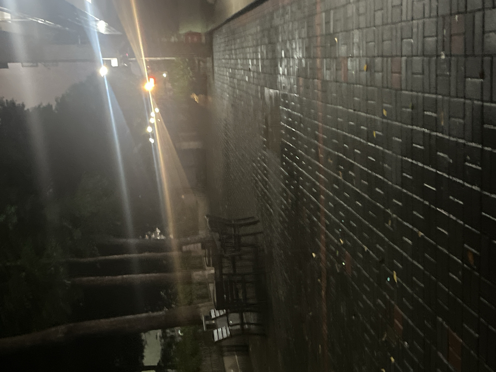 The image displays an outdoor, nighttime scene in Yuzhong District, China. A dark, wet paver pathway occupies the foreground and midground, exhibiting reflective surfaces consistent with recent rainfall. The pavers are arranged in an alternating pattern of dark gray and some lighter, possibly reddish-brown, rectangular units. Scattered fallen leaves are visible on the wet pathway. To the left, several tall, dark tree trunks rise vertically, with their foliage largely obscured by darkness. An unoccupied outdoor seating area, consisting of at least two dark tables and multiple chairs, is positioned near the trees on the left. In the background on the right, a building structure is partially visible. Multiple artificial light sources are present, creating prominent horizontal and diagonal lens flares across the image. Notable light sources include a bright white/blue light on the right and a distinct red light further in the distance. No individuals are present in the scene.