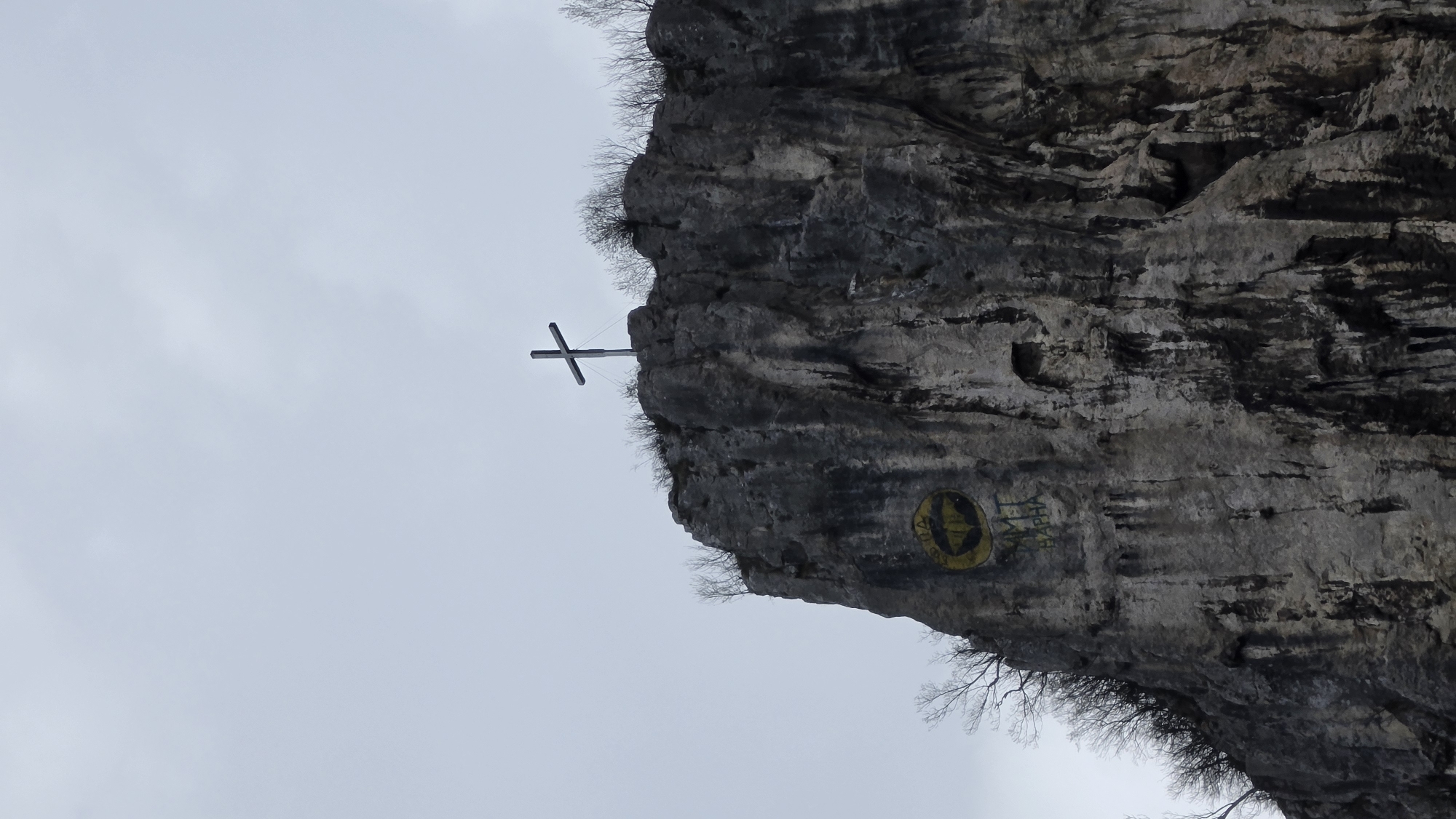 A photograph shows a rock formation with a large metallic Christian cross near the top, set against a cloudy sky. The rock face is gray and textured, with sparse vegetation clinging to its edges. Below the cross, a yellow circular emblem with a stylized symbol is visible on the rock face, accompanied by some faded graffiti. The scene appears to be outdoors, likely a mountainous or elevated area. This landmark is located in Dryanovo, Bulgaria.