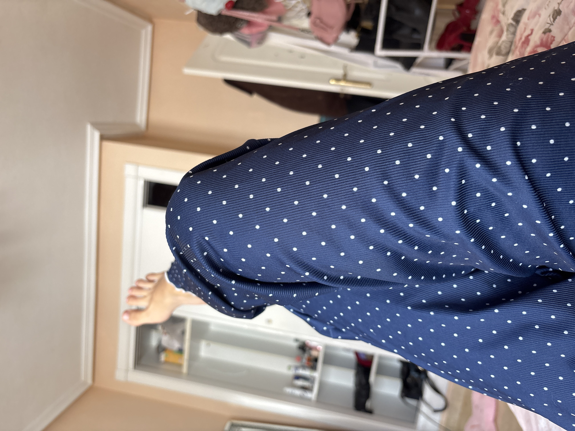 The image shows a close-up view of a person's legs, clad in navy blue polka-dot pajamas, resting on a bed. The bedspread has a floral pattern. In the background, a white wardrobe or closet is visible, containing various items on shelves, including what appear to be bottles and bags. To the right, a closet door with a gold handle is partially open, revealing more items, possibly clothing and accessories, hanging or stored within. The room has pale peach-colored walls and white crown molding. The lighting suggests an indoor environment. The image does not provide any information about the city of Orihuela, Spain.