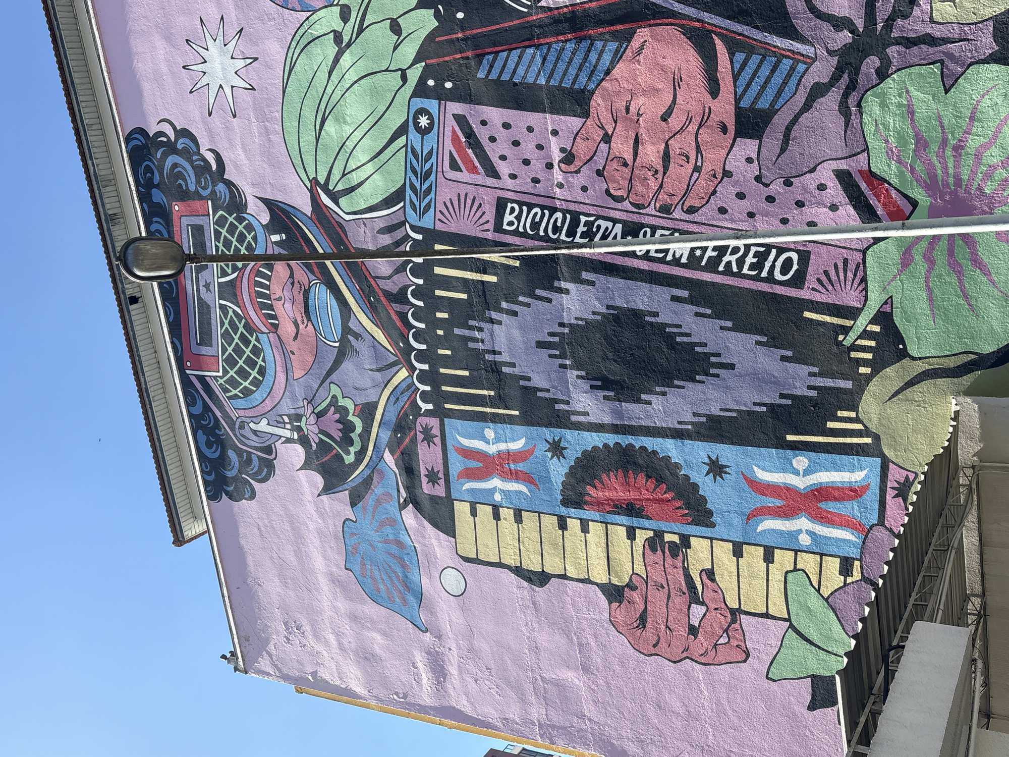 This image captures a vibrant mural painted on the side of a building in São Paulo, Brazil. The mural features a stylized, colorful depiction of a person playing an accordion. The figure has a large, elaborate headdress and is wearing oversized sunglasses. The accordion itself is adorned with geometric patterns and text that appears to read "BICICLETA SEM FREIO." The background of the mural is a soft pink, and the artwork is rendered in bold, graphic lines and bright colors. A street lamp stands in front of the mural, partially obscuring the artwork. The sky above is clear and blue, and the top of a building with a tiled roof is visible. The mural is located on a street, with part of a building and a metal awning visible at the bottom of the frame.