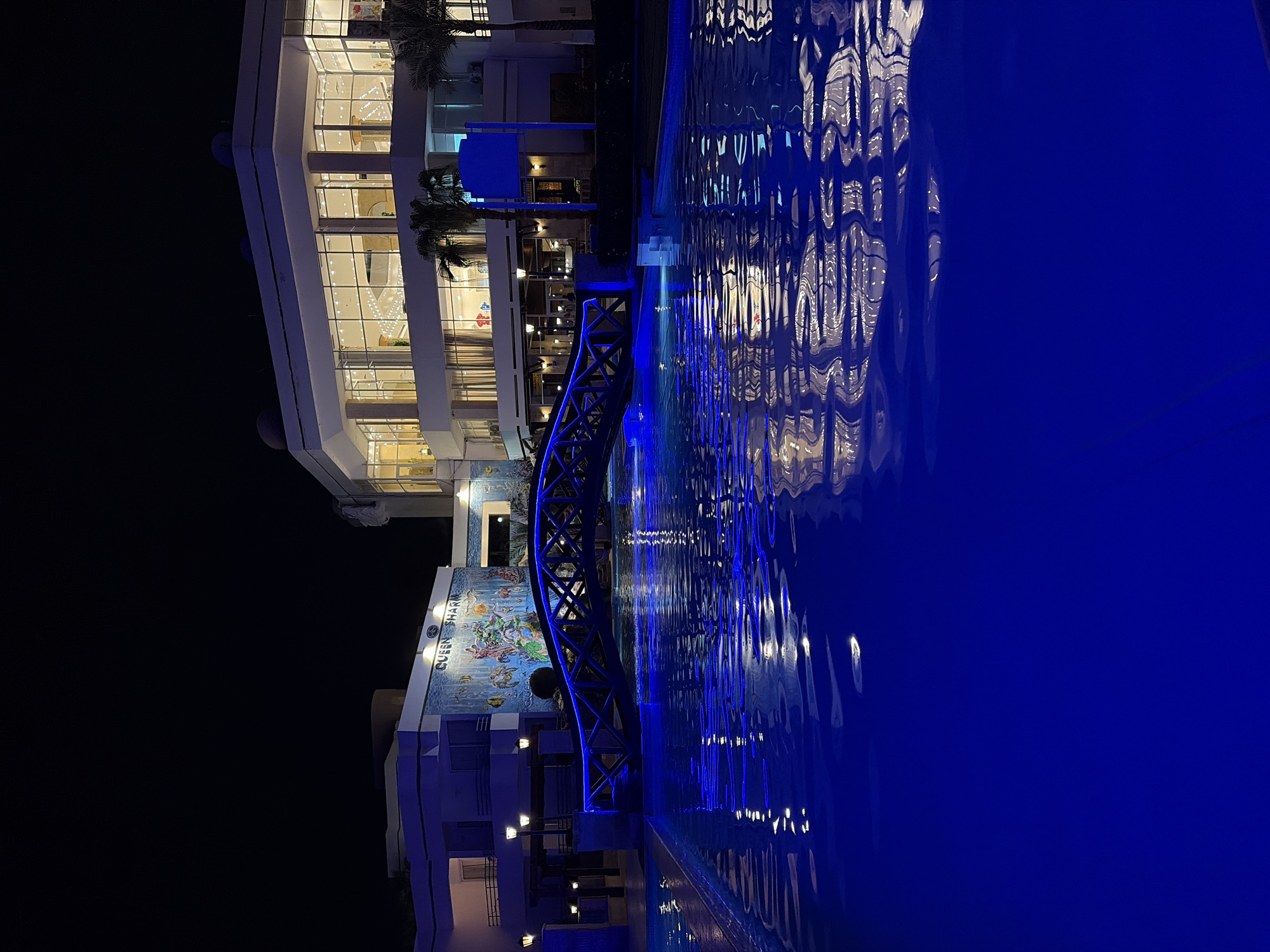 The image depicts a night scene in Sharm El Sheikh, Egypt, showing the "Queen Sharm" hotel. A pedestrian bridge with blue neon lighting arches over a body of water, reflecting the hotel's lights and the bridge's illumination. The hotel structure itself is multi-storied, with illuminated windows. A mural is visible on one section of the building. The surrounding environment is dark, with the lighting primarily concentrated on the hotel, bridge, and water. The water surface is rippled, creating distorted reflections.