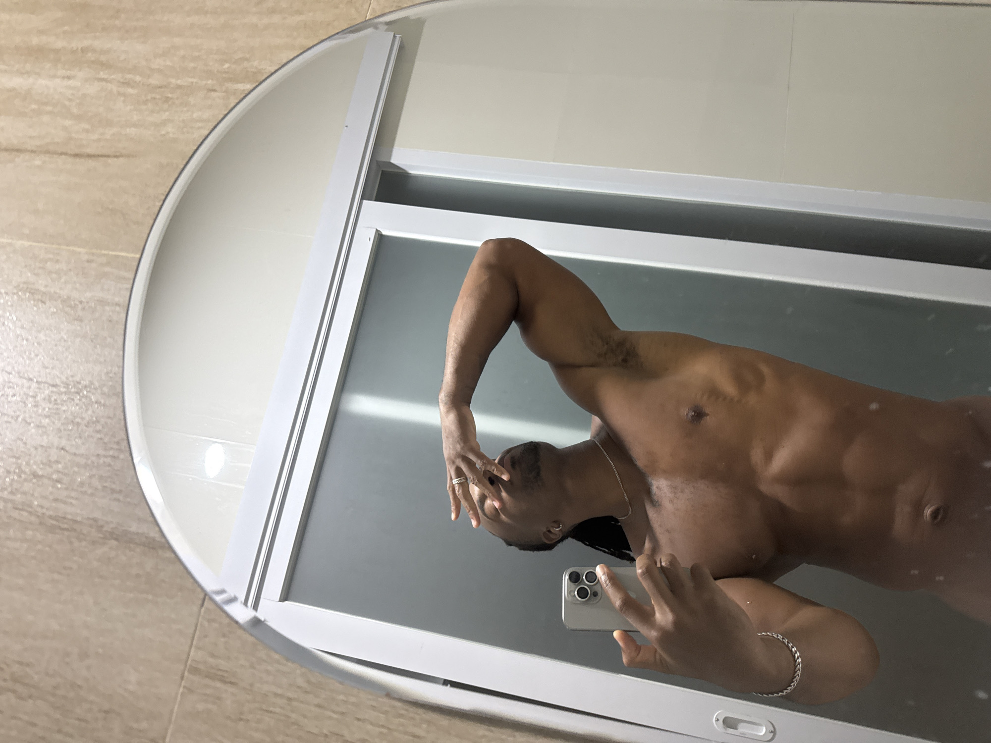 A man is taking a selfie in a mirror. The mirror is an oval shape with a white frame. The man is shirtless and has a muscular physique. He is holding a smartphone in his right hand and his left hand is near his face. He is wearing a silver bracelet on his right wrist and a silver chain around his neck. The mirror is positioned against a white wall and a beige tiled wall. The location is Unknown, Unknown.