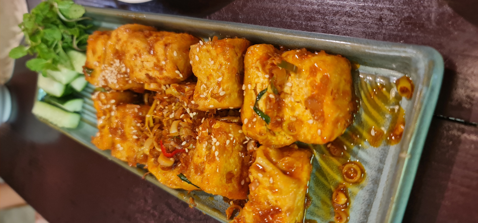 A close-up shot of a rectangular ceramic plate filled with fried tofu, cucumber slices, and garnish, likely taken in a restaurant in Đà Nẵng, Vietnam. The tofu pieces are arranged in a line, glazed with a reddish-orange sauce, and sprinkled with sesame seeds. Some pieces are topped with sauteed vegetables and a thin sliver of red chili. Cucumber slices and sprigs of garnish are situated at the edge of the plate. The plate has a gray-blue border with brown, amber-colored sauce smears. The background shows a blurred dark wooden surface.