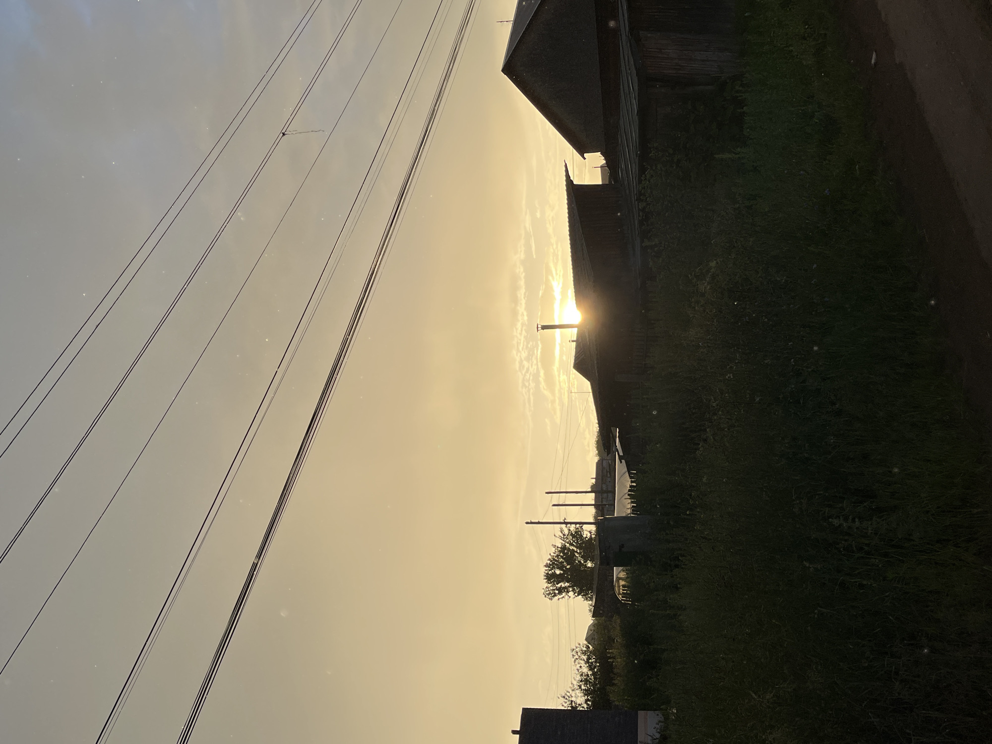 This image depicts a rural landscape during sunset in the village of Olkhovka, Russia. The sky is a pale yellow and orange hue, with scattered clouds. Several power lines are strung across the upper portion of the frame. In the middle ground, there are several low-lying wooden structures with sloped roofs, some appearing to be sheds or outbuildings. A tall chimney protrudes from one of the roofs, with the setting sun partially visible behind it. Dense green foliage and tall grass cover the foreground and the area surrounding the structures, suggesting an overgrown or unkempt environment. To the left, a dark, rectangular structure is partially visible, possibly a building or wall. The overall scene conveys a sense of quiet twilight in a rural setting. There are no people or animals visible in the image.