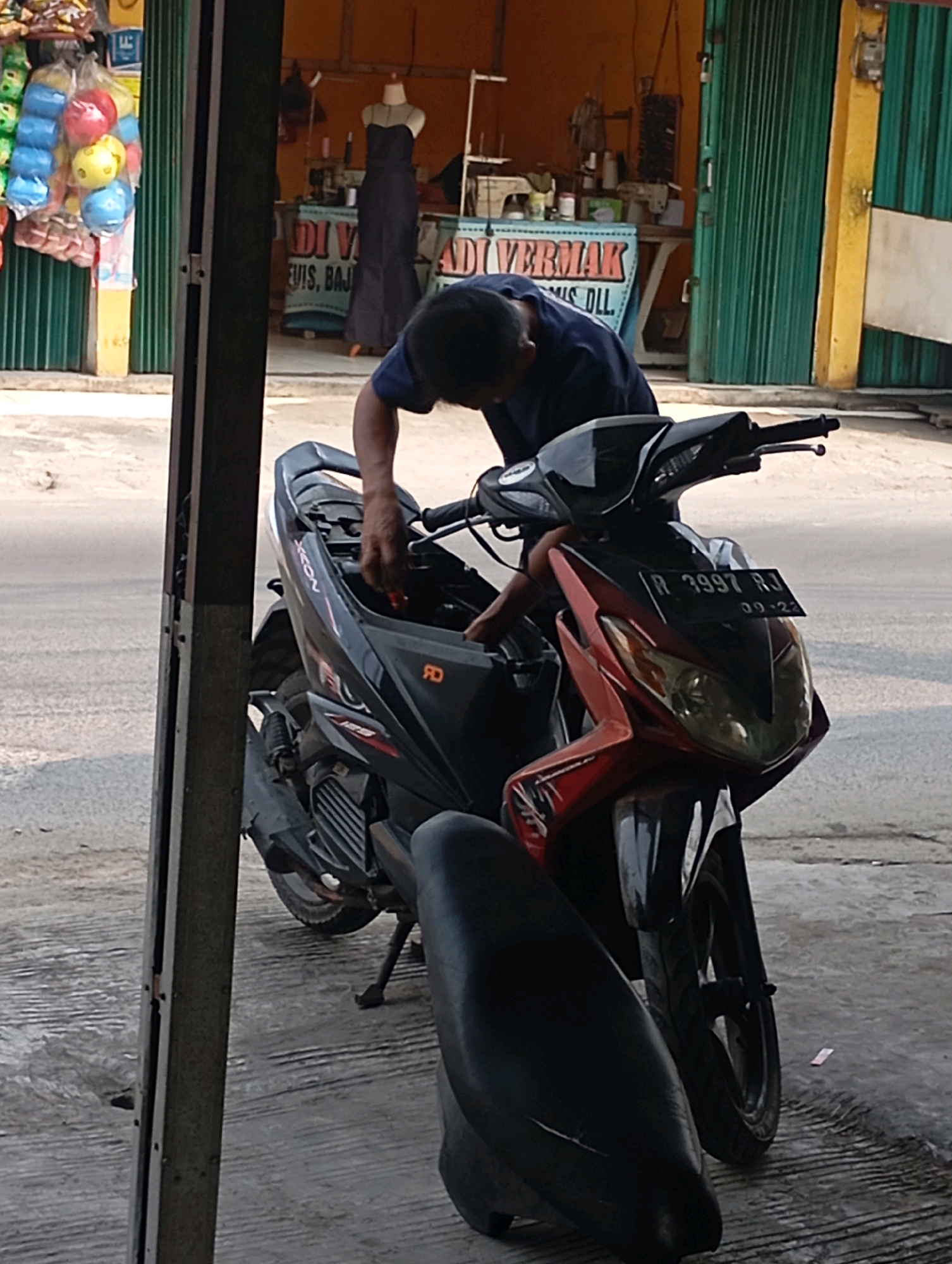 A man, wearing a dark blue t-shirt, is working on a black and red motorcycle in front of a shop in Babelan, Indonesia. The motorcycle's seat is open, and the man is leaning over it, appearing to be performing maintenance or repairs. Behind the motorcycle, the shop has a green metal roll-up door and a sign that reads "ADI VERMAK". To the left of the shop, a display of colorful plastic balls in plastic bags is visible. The ground is paved with concrete.
