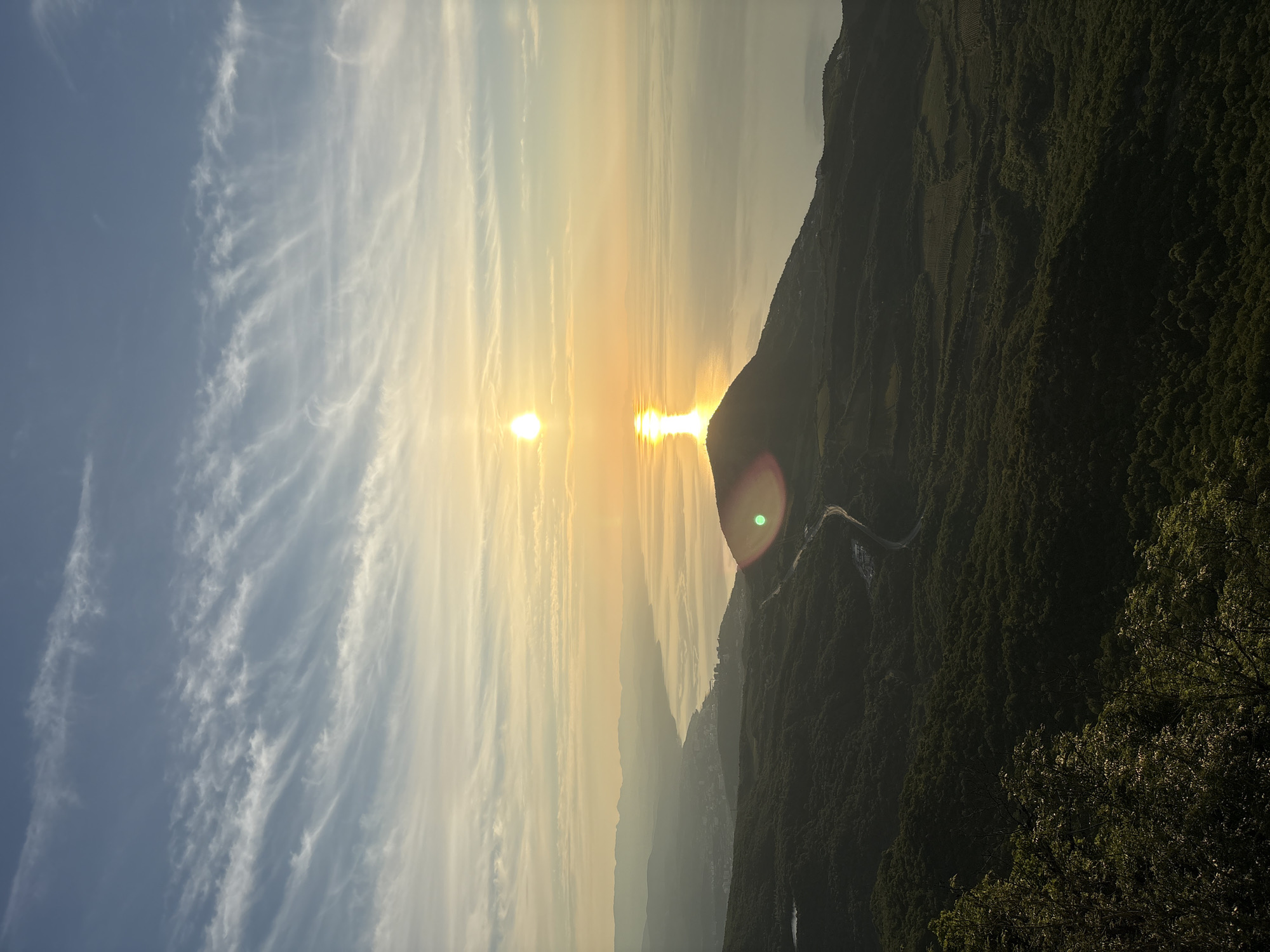This image depicts a coastal and mountainous landscape in Malyi Mayak, Ukraine, observed from a high vantage point during either sunrise or sunset. The sky, occupying the upper third, transitions from light blue at the zenith to pale yellow-orange near the horizon, featuring scattered, elongated, wispy white clouds exhibiting wave-like patterns. The sun, a brilliant yellow-orange orb, is positioned slightly left of the image's vertical center, casting a strong, luminous reflection path vertically down the calm, gray-blue sea.
The sea dominates the middle and right portions of the image, appearing smooth and featureless apart from the sun's reflection and subtle horizontal tonal variations. Along the left side of the image, a curving coastline stretches into the distant haze. A town or city, characterized by numerous buildings and structures, is visible along this coastal strip, backed by receding layers of hazy mountains in the far distance.
The foreground and mid-ground consist of dark green, undulating terrain heavily covered by dense deciduous forest or scrubland. A paved road, appearing as a light gray ribbon, winds through this forested landscape in the mid-right, with some clearings and what appears to be a small commercial or residential complex adjacent to it. A small, dark body of water (possibly a pond or reservoir) is faintly visible within the forested area in the upper left of the landmass. A prominent circular lens flare with red and green chromatic aberrations is present in the lower right quadrant, obscuring part of the forested hill. No individuals or specific actions are discernible within the frame.