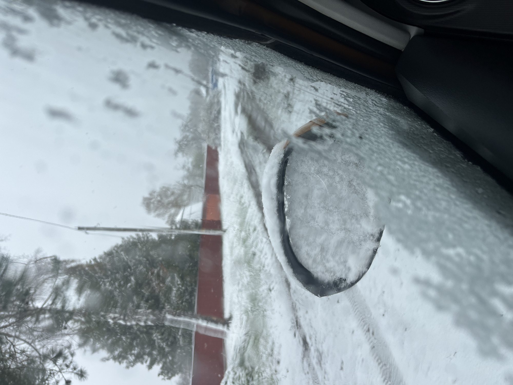 The image depicts a snow-covered scene viewed from inside a vehicle, likely a car. The side mirror is heavily coated in snow and ice. Outside the window, trees and a red building are visible, also covered in snow. Tire tracks can be seen in the snow-covered ground. The location is presumably Раздолье, Russia. The weather conditions suggest a winter season with significant snowfall. There are no people visible in the image.