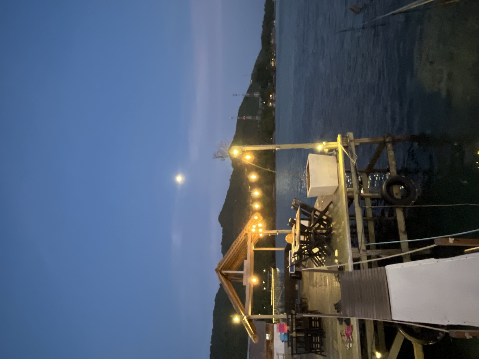 The image depicts a scenic view of a waterfront restaurant at dusk in Karimunjawa, Indonesia. The restaurant, built on a wooden pier extending into the calm, dark blue water, features a thatched-roof gazebo over a seating area. String lights, glowing yellow, are draped across the gazebo and along the pier, illuminating the tables and chairs set up for dining. In the foreground, a ramp leads down to the pier. The water reflects the lights and the twilight sky. In the background, lush green hills rise, dotted with structures and communication towers. A bright full moon hangs in the clear, deep blue sky. There are no people visible in the frame.