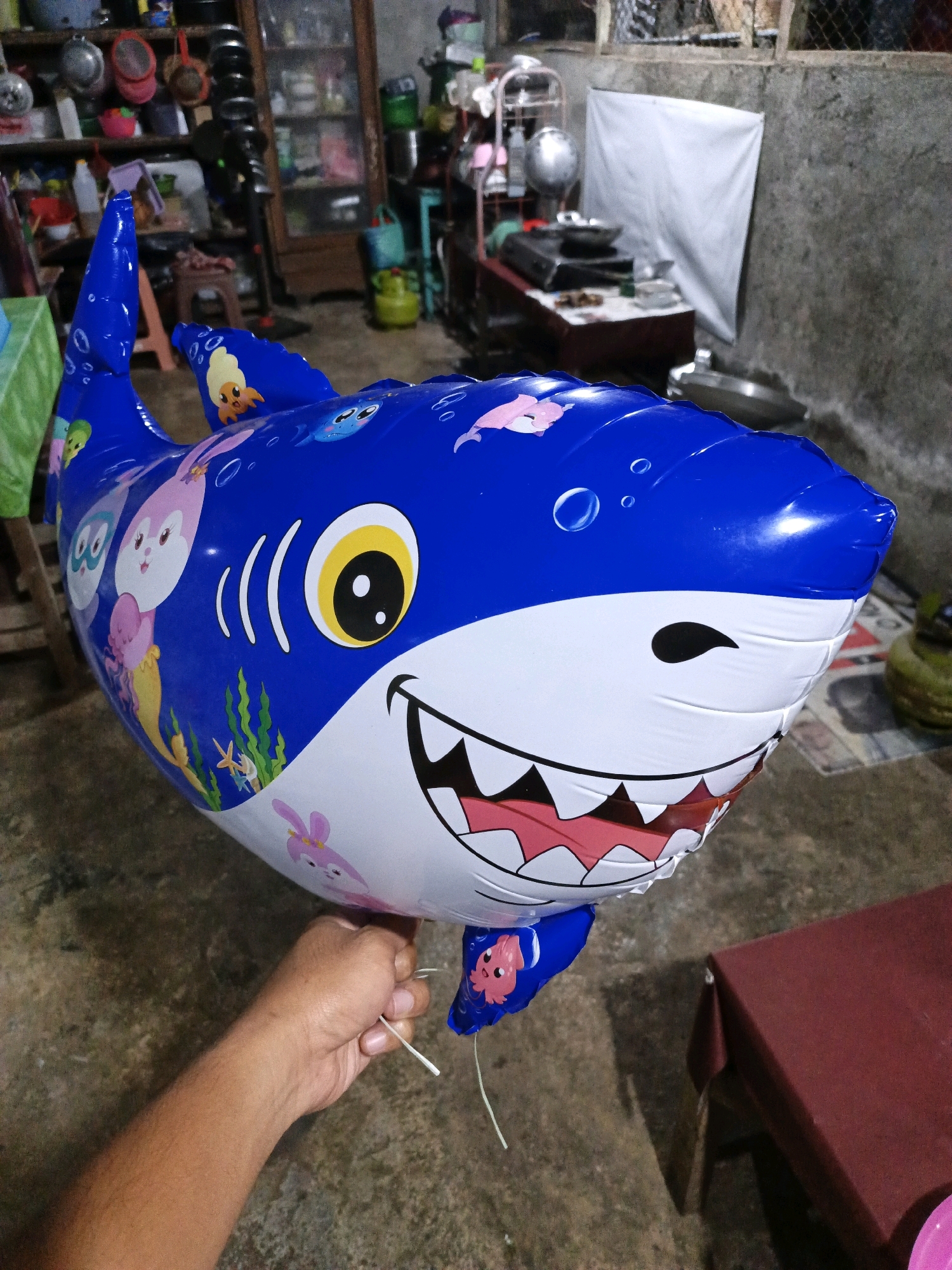 In Keleyan, Indonesia, a blue inflatable shark balloon with cartoon characters on its surface is being held by a person's right hand. The background suggests an outdoor setting or an open-air structure, with a concrete floor and a textured wall. In the distance, shelves are visible, filled with various items including pots, pans, and containers. A red tablecloth covers a table on the right side of the frame, and a white sheet is draped over an object in the background. The lighting suggests it is daytime, with some shadows cast on the ground.