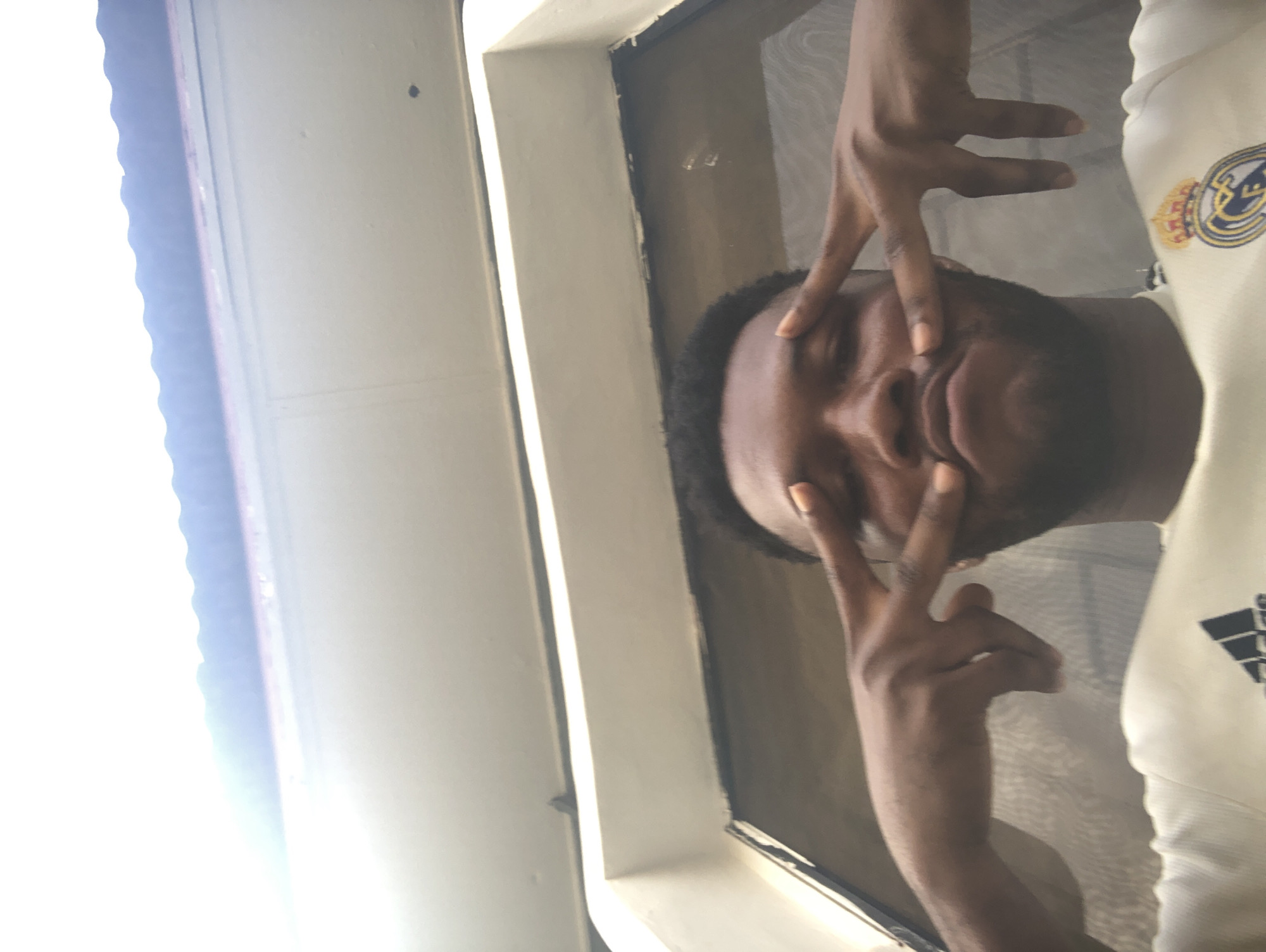 A person with dark skin and short black hair is looking down at the camera with their hands positioned making a V shape over their eyes. The person is wearing a white shirt with the Real Madrid logo and Adidas stripes. The person is standing inside a room near a window with a white frame and white walls. The location is Unknown, Unknown.