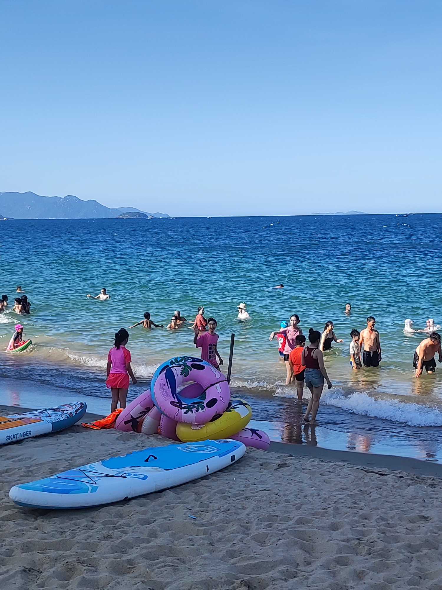This is a vertical, eye-level, full-shot photograph taken on a sunny day at a beach in Nha Trang Ward, Vietnam. The scene captures a lively beach environment with a clear blue sky above and calm, turquoise ocean water. In the foreground, two paddleboards rest on the sandy beach, one labeled "SKATINGER." A collection of inflatable swimming rings, predominantly pink and yellow, are piled near the water's edge.
The ocean is populated with numerous people, including adults and children, engaging in various water activities. Some are wading in the shallow water, while others are swimming or floating further out. The water is clear enough to see some of the people's legs and the sandy bottom near the shore.
The background features a hazy, mountainous coastline, a common characteristic of Nha Trang. The overall atmosphere is one of recreation and enjoyment, with people seemingly relaxed and having fun in the warm weather. The clear sky and bright sunlight suggest it is daytime, likely midday or early afternoon.