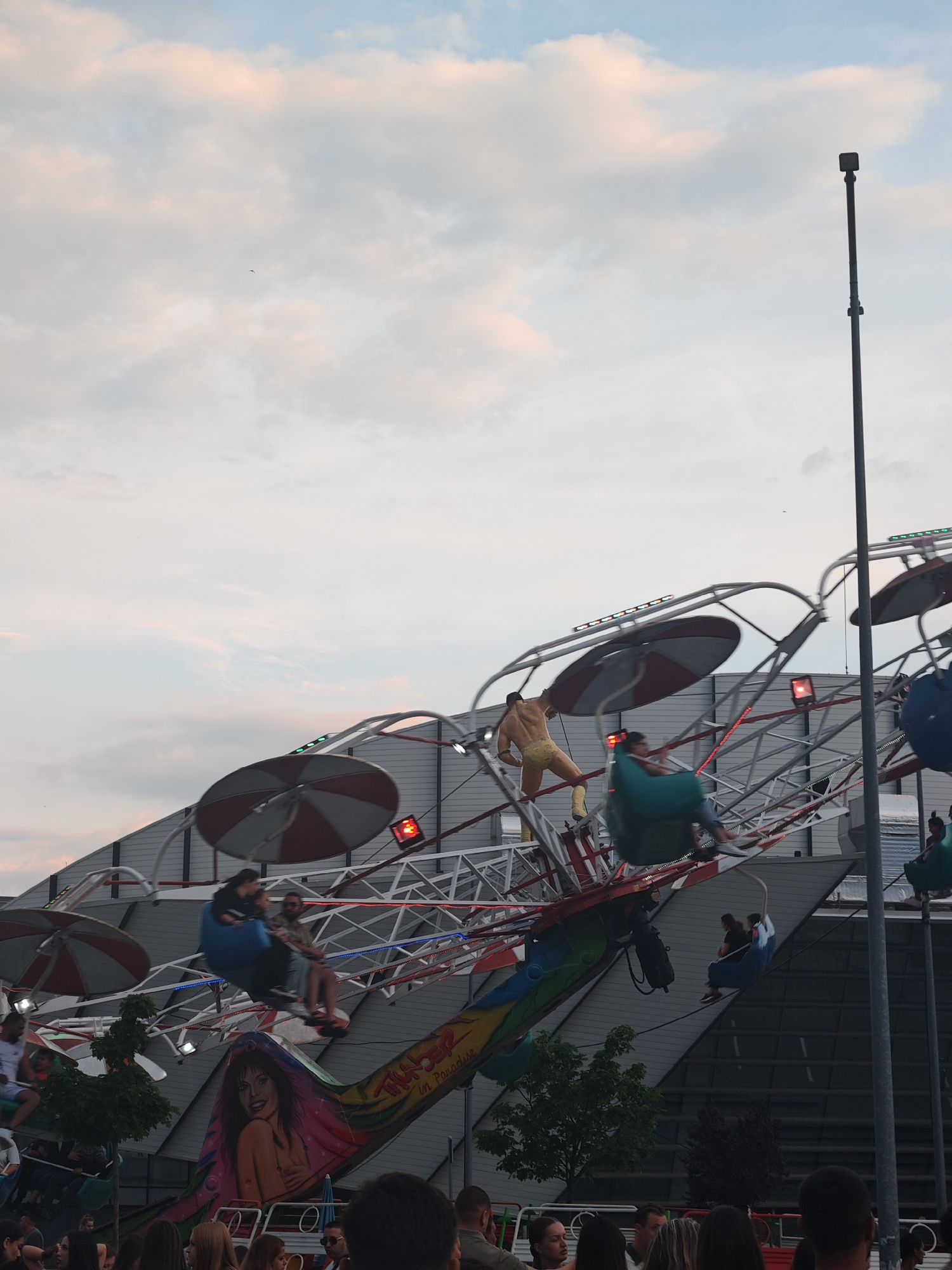 The image shows a fairground ride in motion. The ride has several seats, each occupied by one or two people, rotating around a central axis. There is a muscular figure standing on the ride. The ride is decorated with colorful graphics and lights. In the background is a light gray building. The scene is located in the city of Shumen, Bulgaria. There are people standing at the bottom of the frame.