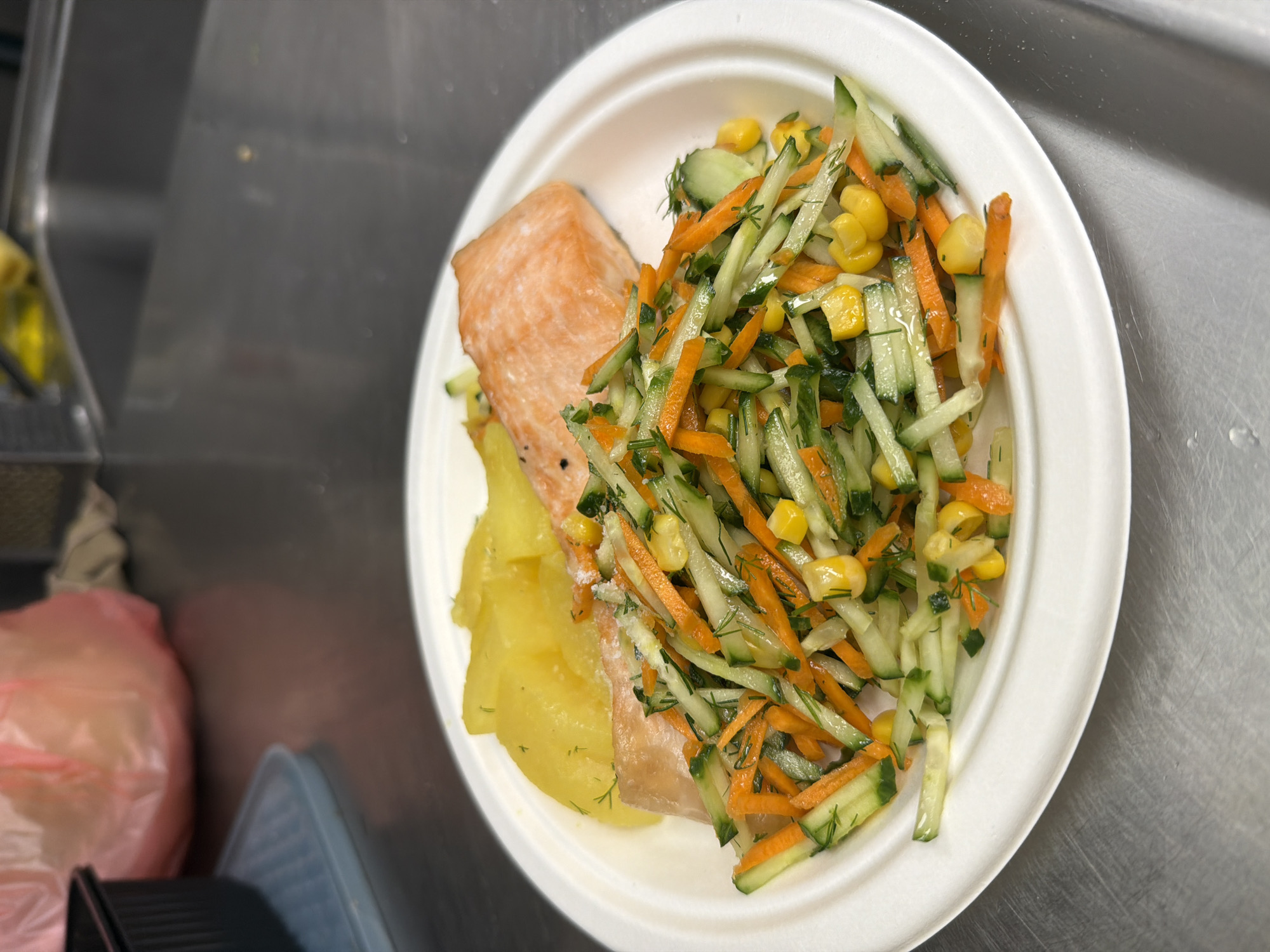 A close-up, eye-level shot shows a white plate of food on a metallic surface. The plate contains a fillet of cooked salmon, a portion of sliced yellow potatoes, and a salad. The salad is composed of julienned cucumbers and carrots, with visible corn kernels and chopped dill. The background is blurred, showing a pink plastic bag and part of a sink area, suggesting a kitchen or food preparation environment. No people are visible in the frame. The lighting appears to be overhead and diffused. The location is not discernible from the image itself.