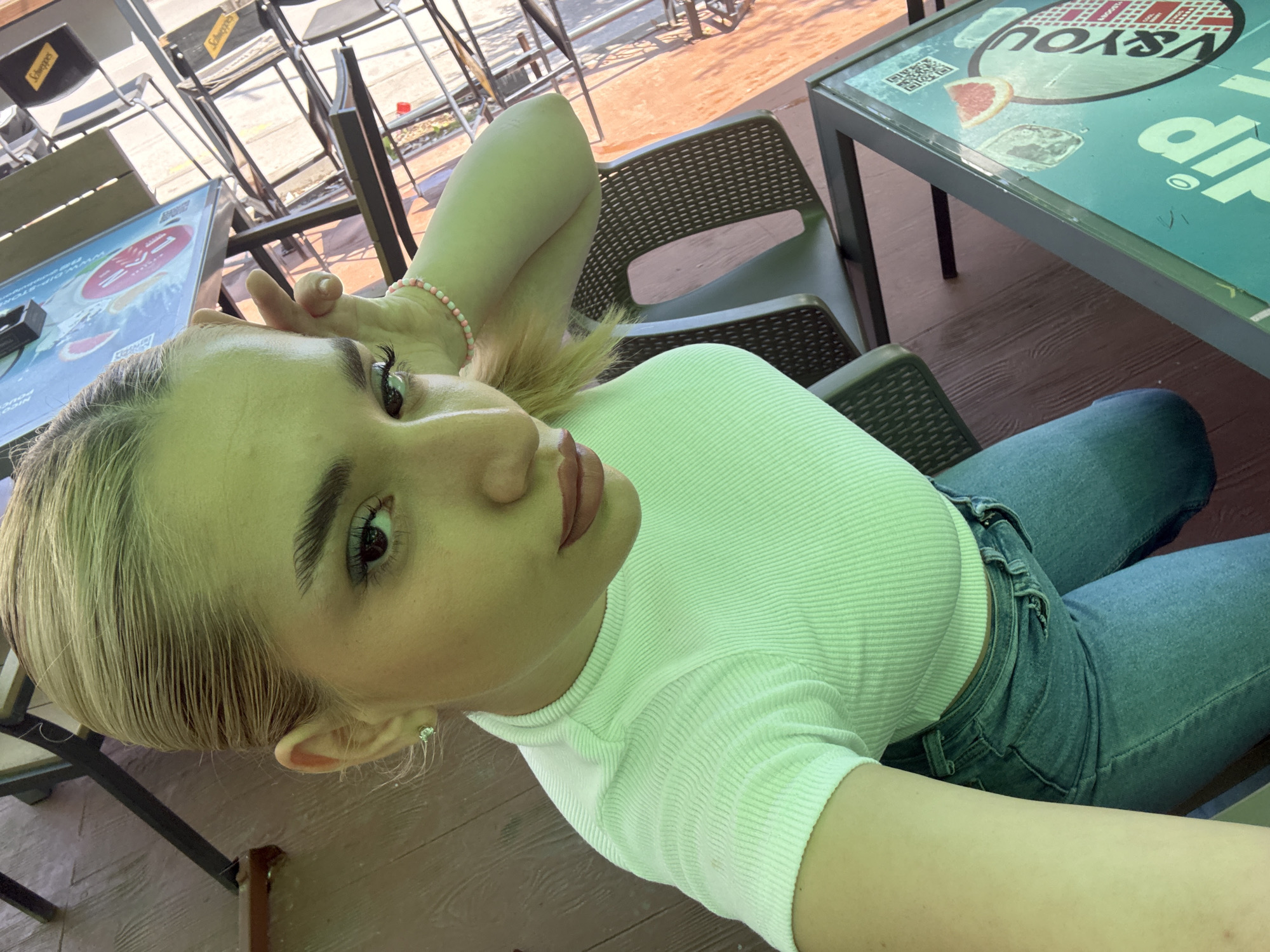 A selfie portrait captures a young woman seated outdoors at what appears to be a cafe or restaurant in Sofia, Bulgaria. The image is taken at a tilted angle, focusing primarily on her face and upper body. She has light blonde or ash-blonde hair, styled smoothly, and is wearing a form-fitting, light green ribbed short-sleeved top paired with blue denim jeans. A delicate bracelet adorned with small pink and white beads is visible on her left wrist, and she wears a subtle stud earring in her visible right ear. Her makeup includes visible eyeliner, mascara, and a natural lip color, as she looks directly at the camera with a neutral expression.
The background shows an outdoor seating area furnished with dark-framed tables and chairs. Some chair backs feature the brand "Schweppes" in white text on black fabric. A table in the foreground has a distinctive green surface displaying graphics such as a slice of grapefruit, a QR code, and inverted text including "V & YOU" and a partially visible "D'YOU." The flooring beneath the seating area is made of wooden planks. The bright, natural illumination suggests the photograph was taken during the daytime, likely under sunny conditions.