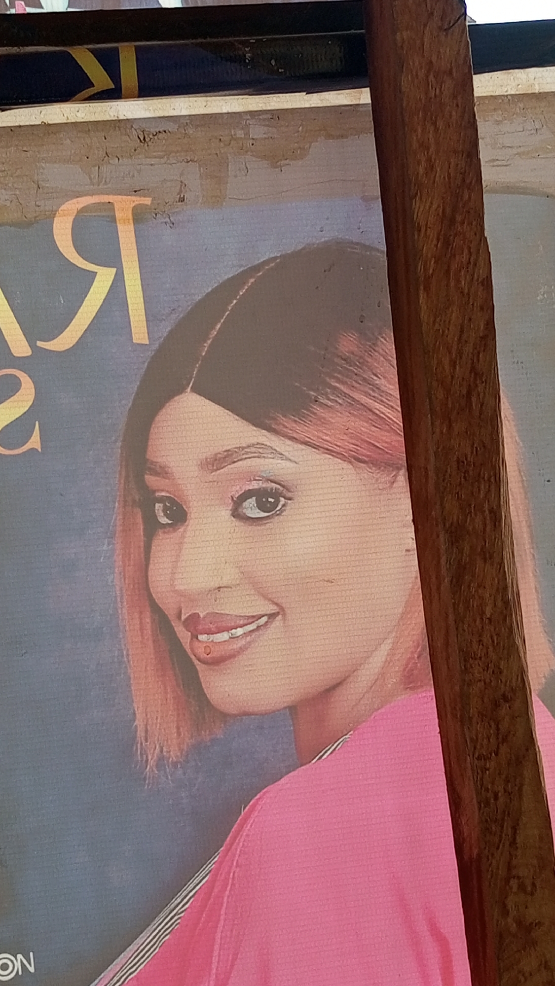 A medium-shot photograph, taken at an upward angle, features a poster of a woman with fair skin, brown hair with pink highlights, and wearing a pink shirt. She is smiling and looking to the left. The poster is mounted on a wooden structure, as evidenced by the weathered wood and wooden frame visible on the right side of the image. A yellow, Cyrillic-like letter "Я" is visible on the upper left of the poster, along with some text below. The poster is mounted on a structure, likely in Sabon Tasha, Nigeria.