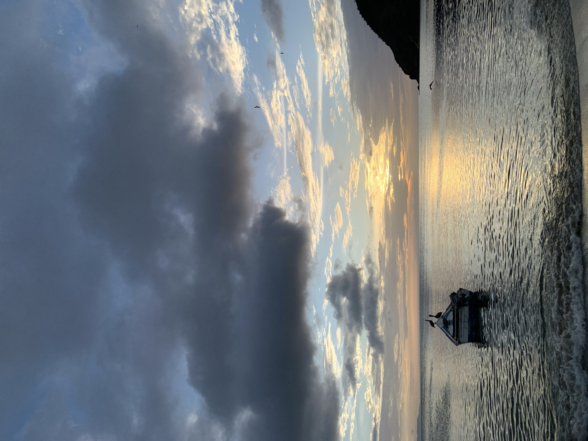 A wide shot captures a serene coastal scene at dusk in Juan Griego, Venezuela. The sky is a dramatic canvas of dark, heavy clouds on the left, transitioning to lighter, wispy formations painted with hues of orange, yellow, and blue as they approach the horizon. The sun, a bright orb, is partially obscured by clouds, casting a golden, shimmering reflection on the calm, dark water.
In the middle ground, a small, weathered boat floats gently on the water. It appears to be made of wood with white and blue paint, and an outboard motor is visible at the stern. Two pelicans are perched atop the boat's structure, silhouetted against the sky. The foreground shows the edge of a sandy beach with small, gentle waves lapping at the shore.
To the right, a dark, silhouetted headland juts out into the sea, its form softened by the fading light. Several birds are visible in the sky, appearing as small dark specks against the colorful clouds. The overall atmosphere is one of peaceful tranquility and the quiet beauty of nature at the close of day.