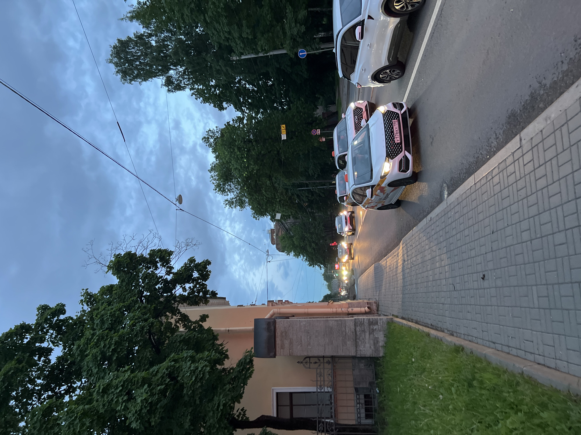 The image depicts a street scene in Saint Petersburg, Russia, during the twilight hours. The perspective is from a sidewalk looking towards a street filled with vehicles. On the left, a building with light-colored walls and a window is visible, bordered by a green grassy area and a paved sidewalk. The street itself is busy with several cars, many of which appear to be taxis, indicated by markings on their roofs and doors. Traffic lights are on, and car headlights illuminate the scene. Trees line the street on the right, and utility wires are strung overhead, suggesting an urban environment. The sky is overcast with a mix of blue and gray clouds.