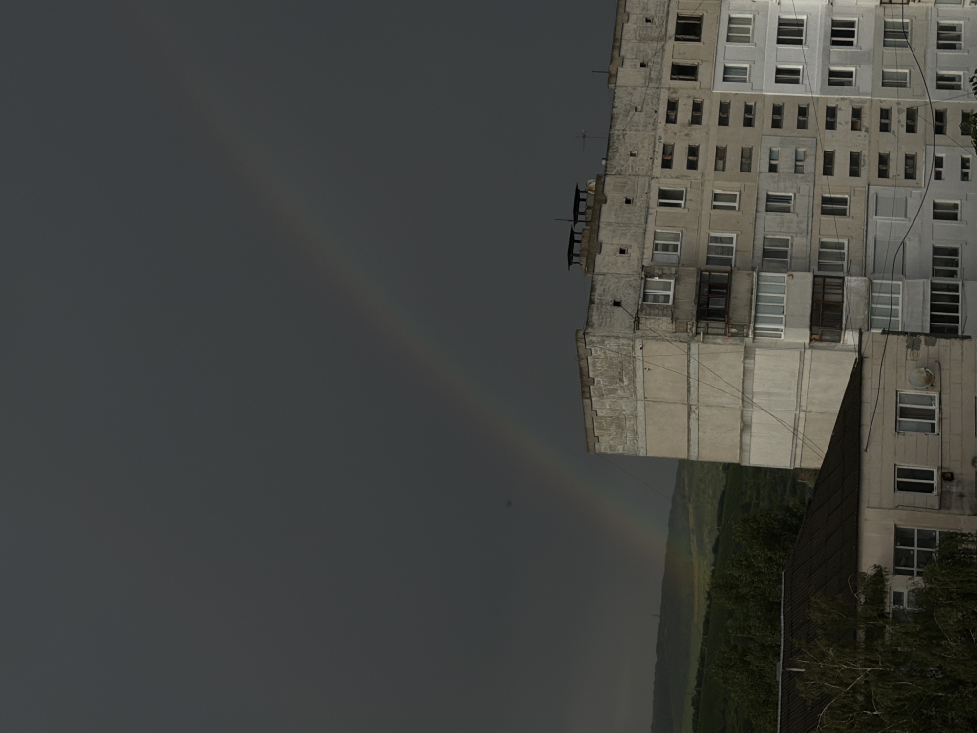 This image depicts a residential building and a smaller structure in the city of Strășeni, Moldova, under a dark, overcast sky with a faint rainbow visible. The main subject is a tall, multi-story concrete apartment building with numerous windows. To the left of this building, a lower, darker-roofed structure is partially visible. In the background, a lush green hillside can be seen, suggesting a semi-urban or suburban environment. The overall lighting is dim, consistent with pre-storm or post-storm conditions. There are no people or animals present in the frame. The focus is on the architecture and the atmospheric conditions.