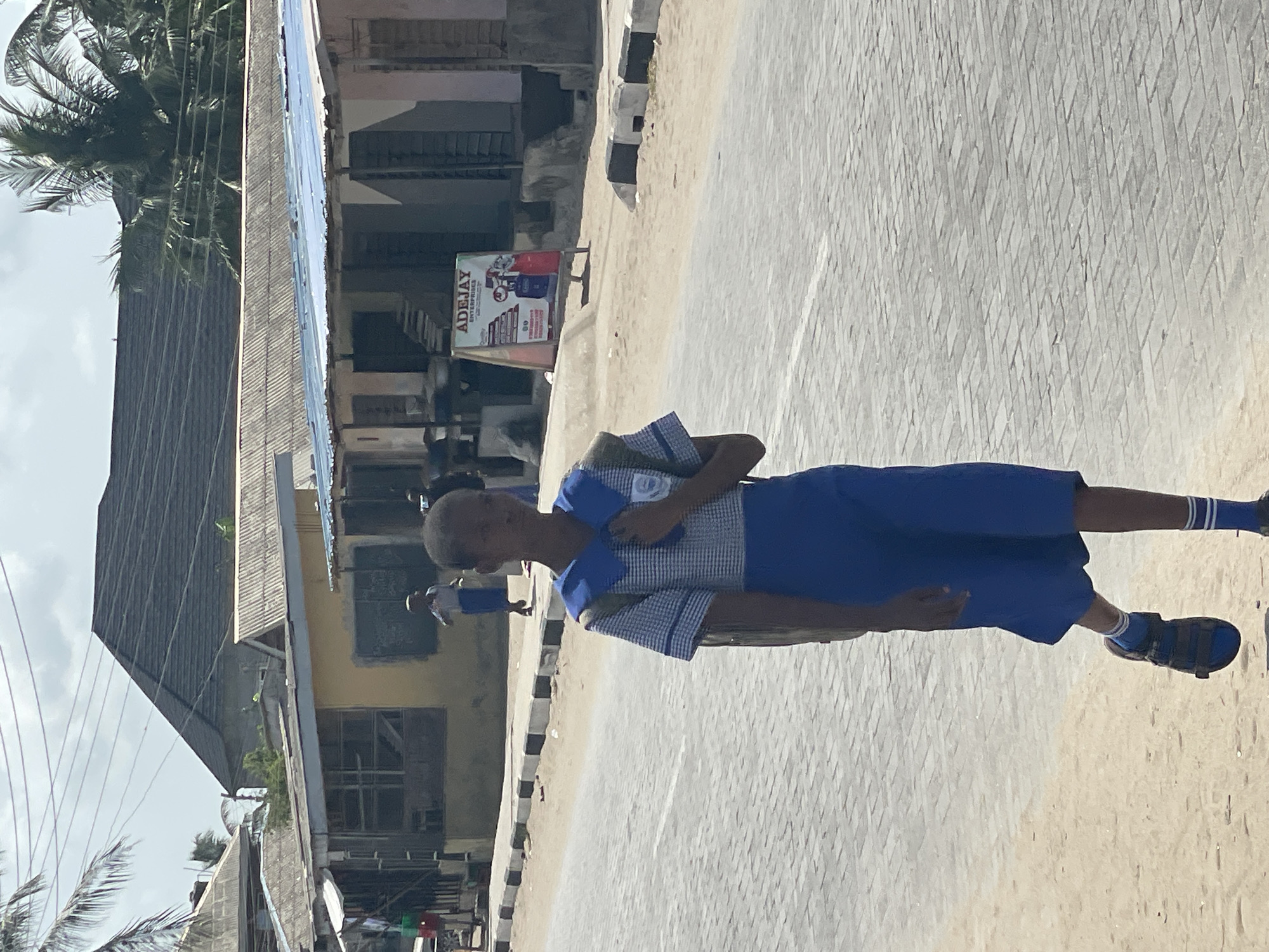 A young person wearing a blue and white uniform walks down a paved street in Badagry, Nigeria. The street is lined with buildings, including one with an "ADEJAY" sign. Palm trees are visible in the background. The individual carries a bag or books. The sky is overcast. The street surface has a diamond-shaped pattern.