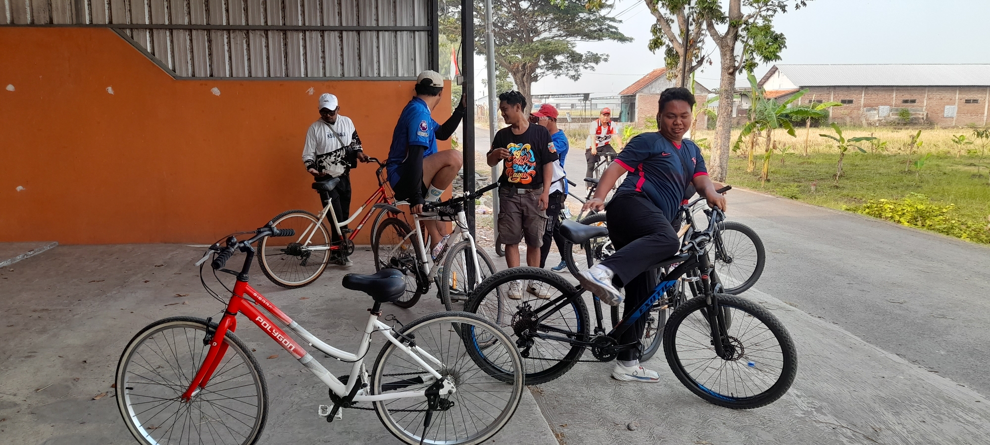 In Loning, Indonesia, a group of seven people are gathered outdoors near a paved road and an orange building with a corrugated metal roof. Several bicycles are present, with five of them in the foreground. The people appear to be of varying ages and are dressed in casual attire, with some wearing athletic clothing. One individual, wearing a blue and red t-shirt and black pants, is seated on a black bicycle, leaning to the right with their left foot extended. Another person, in a black t-shirt with colorful lettering and brown shorts, stands to the left, looking towards the seated individual. Two people in the background are also standing near bicycles, one in a white long-sleeved shirt and the other in a blue long-sleeved shirt. Two additional individuals are partially visible in the background. The environment includes trees, a field with dry grass, and a brick building in the distance. The lighting suggests it is daytime, with clear skies.