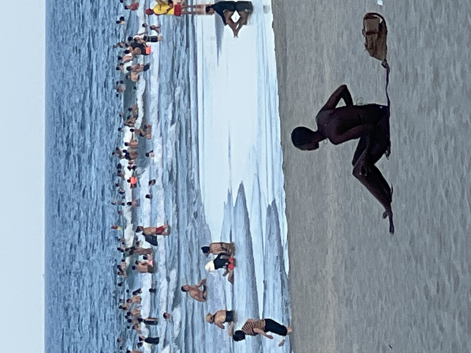 A crowded beach in Da Nang, Vietnam, shows people swimming and sunbathing. In the foreground, a dark-skinned individual sits on the wet sand facing left, with a duffel bag to their right. In the middle ground, several people are wading in the shallow water. The background is filled with numerous individuals swimming and playing in the ocean, with waves breaking. The sky is overcast.