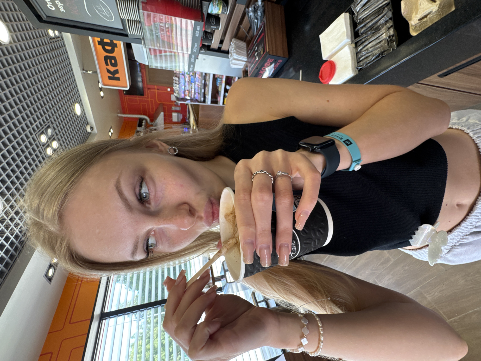 A young Caucasian female with blonde hair and light skin is holding a disposable coffee cup in her right hand and a wooden stir stick in her left hand, which she is using to stir the coffee. She is wearing a black tank top, light gray sweatpants, and several pieces of jewelry, including bracelets on her left wrist and rings on her right hand. A watch is visible on her left wrist, and a turquoise band with writing is around her right forearm. The background suggests an indoor setting, possibly a cafe or convenience store, with shelves stocked with various items and signage in Russian visible. The overall lighting is bright, indicating daytime. There is no specific information to pinpoint the location as Marchugi, Russia.