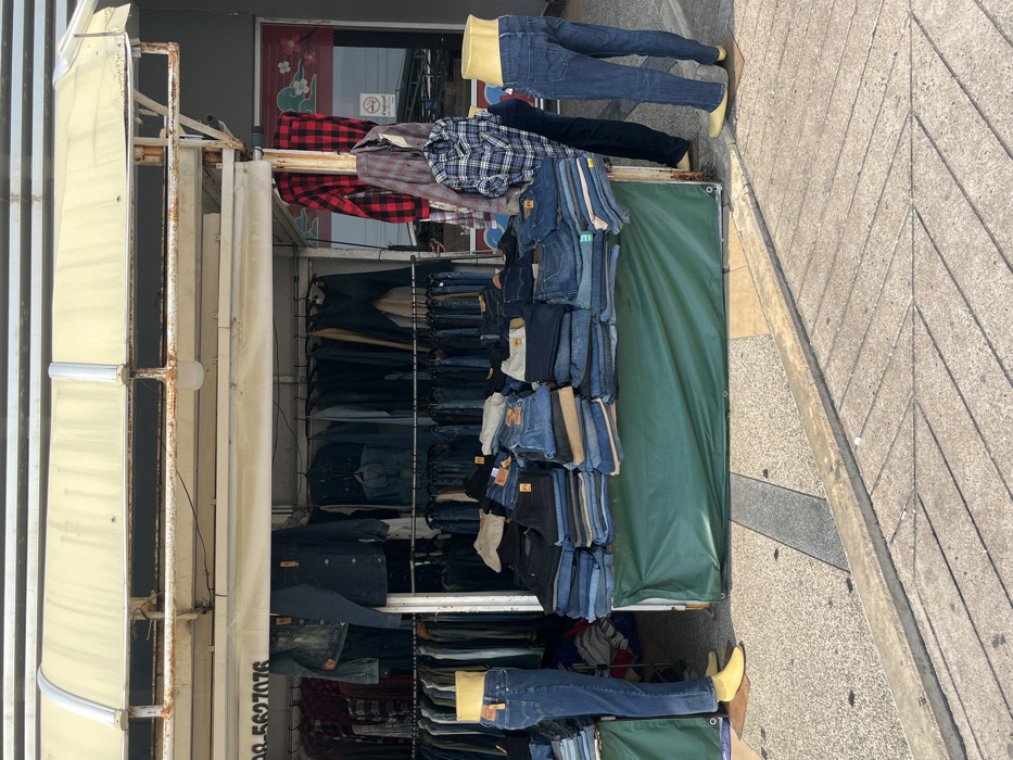 A roadside clothing stall is depicted on a paved outdoor surface in Bang Pu Subdistrict Municipality, Thailand. The stall features a rusty metal frame supporting a light-colored, translucent corrugated awning. Beneath the awning, a large assortment of denim jeans, jackets, and shirts is displayed. Numerous jeans are neatly stacked in piles on a table covered with a dark green tarp, some bearing yellow price tags. Additional jeans and various shirts, including red-and-black and blue-and-white plaid patterns, hang from multiple metal racks behind and above the display table. No living individuals are visible. Two headless, skin-tone mannequins, one legless torso on the left and a full-leg mannequin on the right, both wearing blue jeans, are positioned near the display. Their feet are stylized in yellow. A phone number, '099 562 7076', is partially visible on a sign above the racks. In the background, a modern building with a grey facade features a window adorned with a red and white decorative pattern and a 'No Smoking' sign in Thai script. The ground consists of light-colored paved sections interspersed with darker patterned areas and a concrete curb.